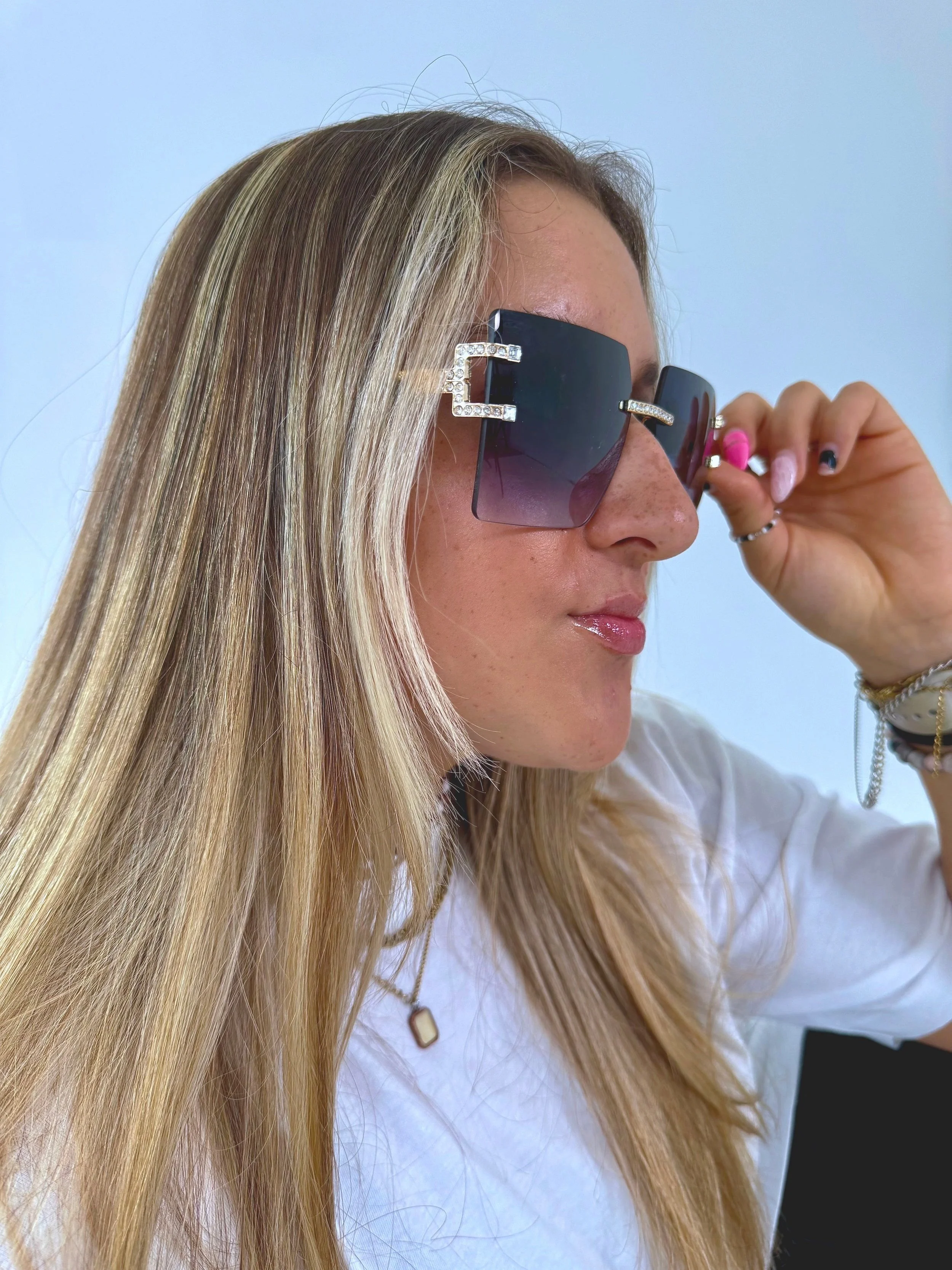 A woman with long blonde hair wearing large, black designer sunglasses with rhinestone details on the hinges, adjusting them with her right hand.