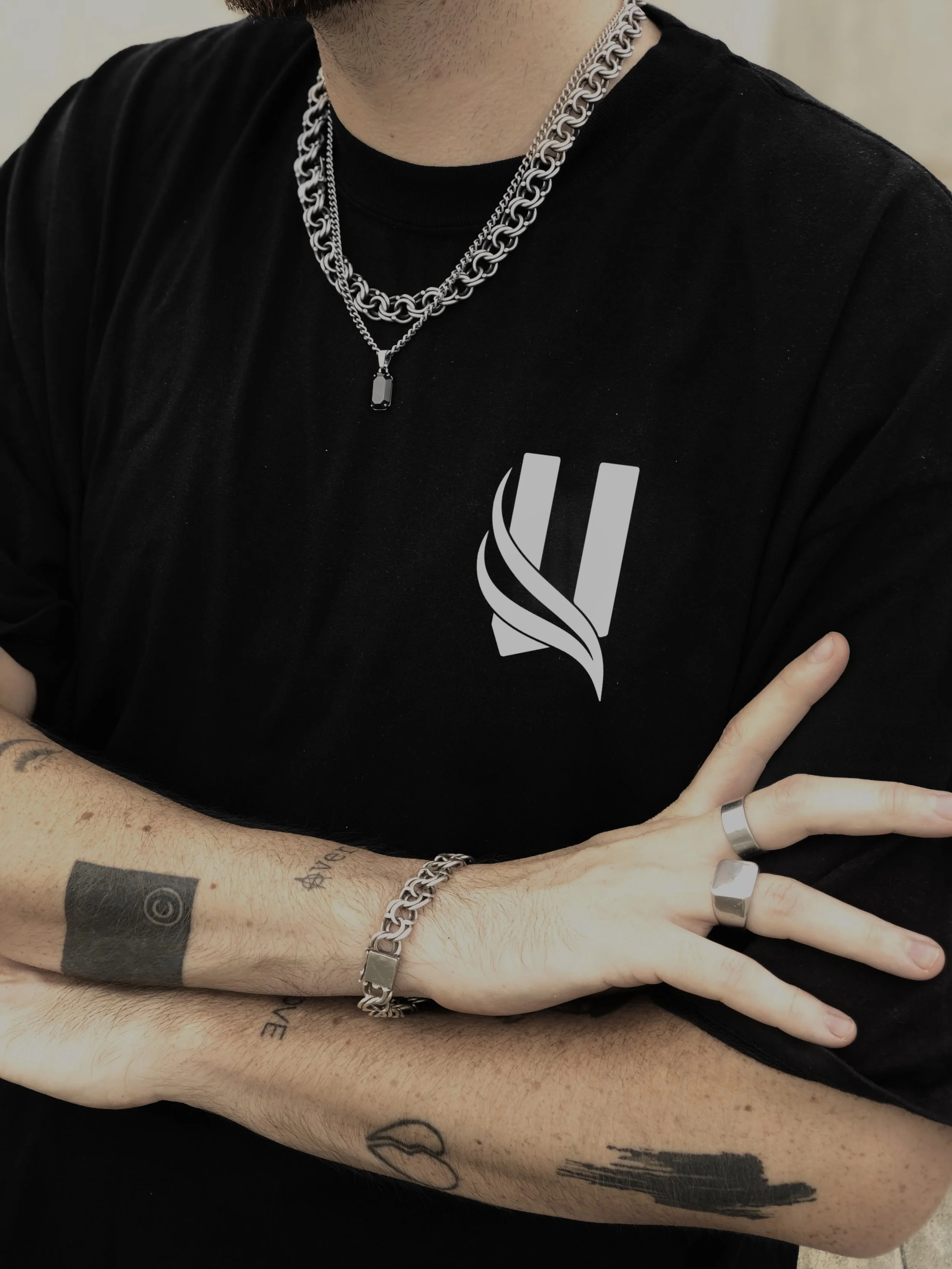Close-up of a person's upper body showcasing layered silver chains around their neck, a black shirt with a white graphic logo, and tattoos on their forearm. They are wearing a ring on the middle finger of their right hand and a bracelet on their left