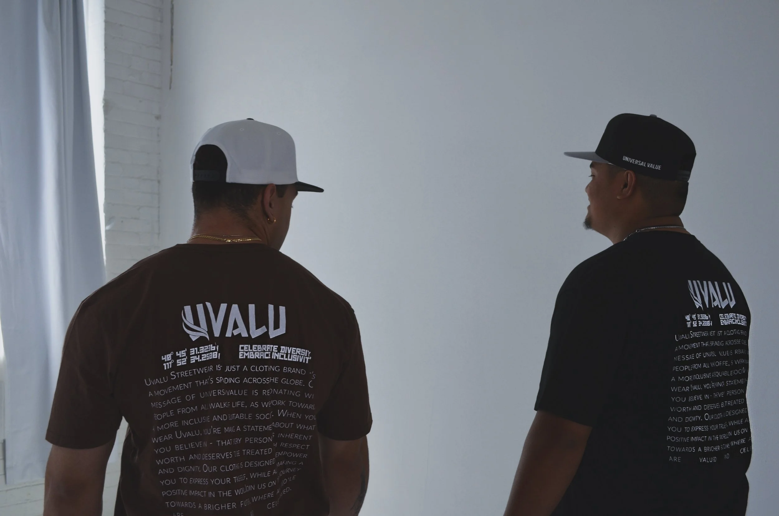 Two men facing away from the camera, standing indoors against a plain white wall. They are wearing black and white caps and black T-shirts with white text on the back that says 'UALU' and other smaller text.