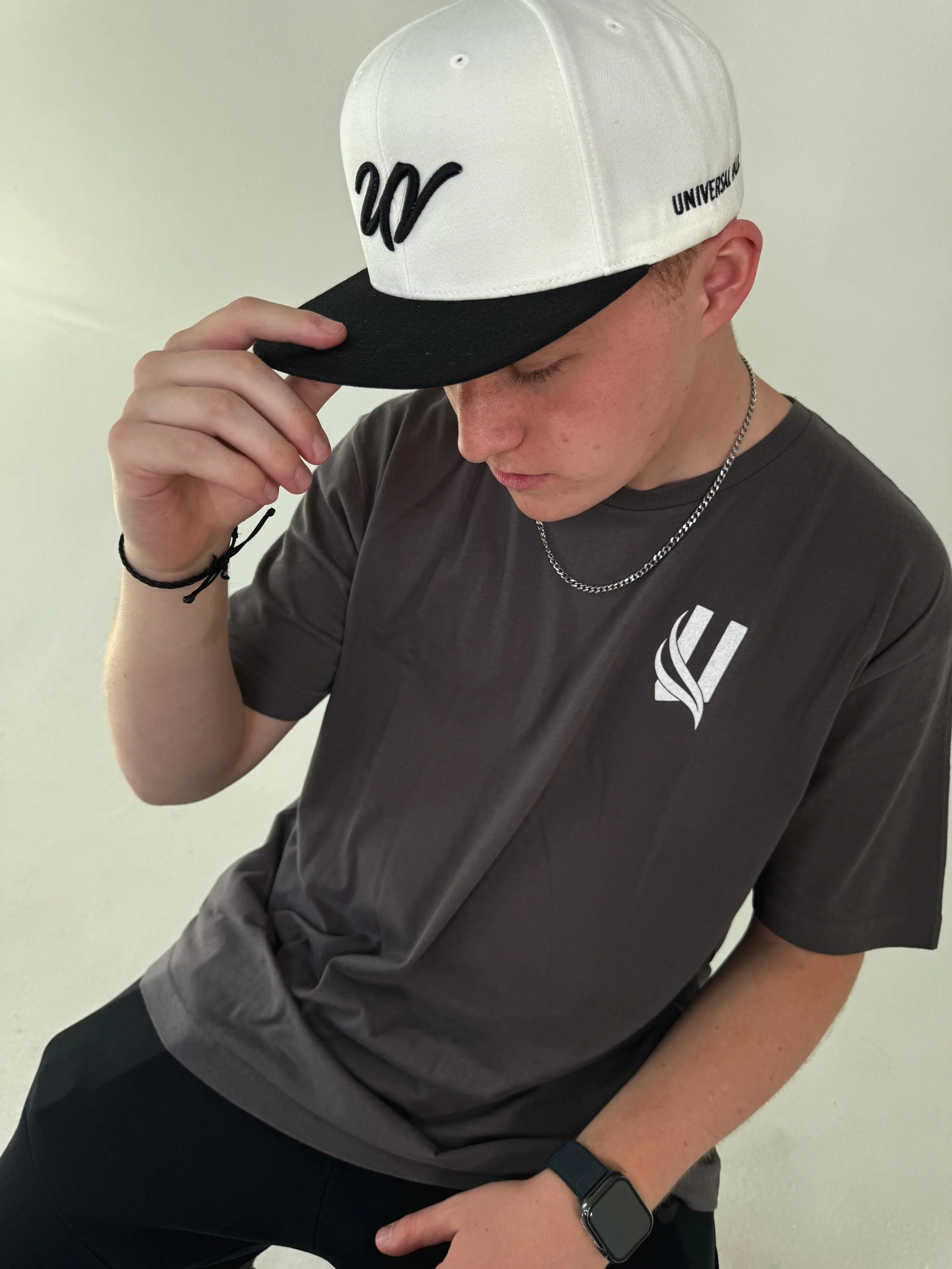 A young man wearing a gray T-shirt with a white logo, a cap with the words '20/20' on the front and 'UNIVERSITY' on the side, a silver chain necklace, black watch, and a black bracelet. He is tilting his head down and holding the brim of his cap with