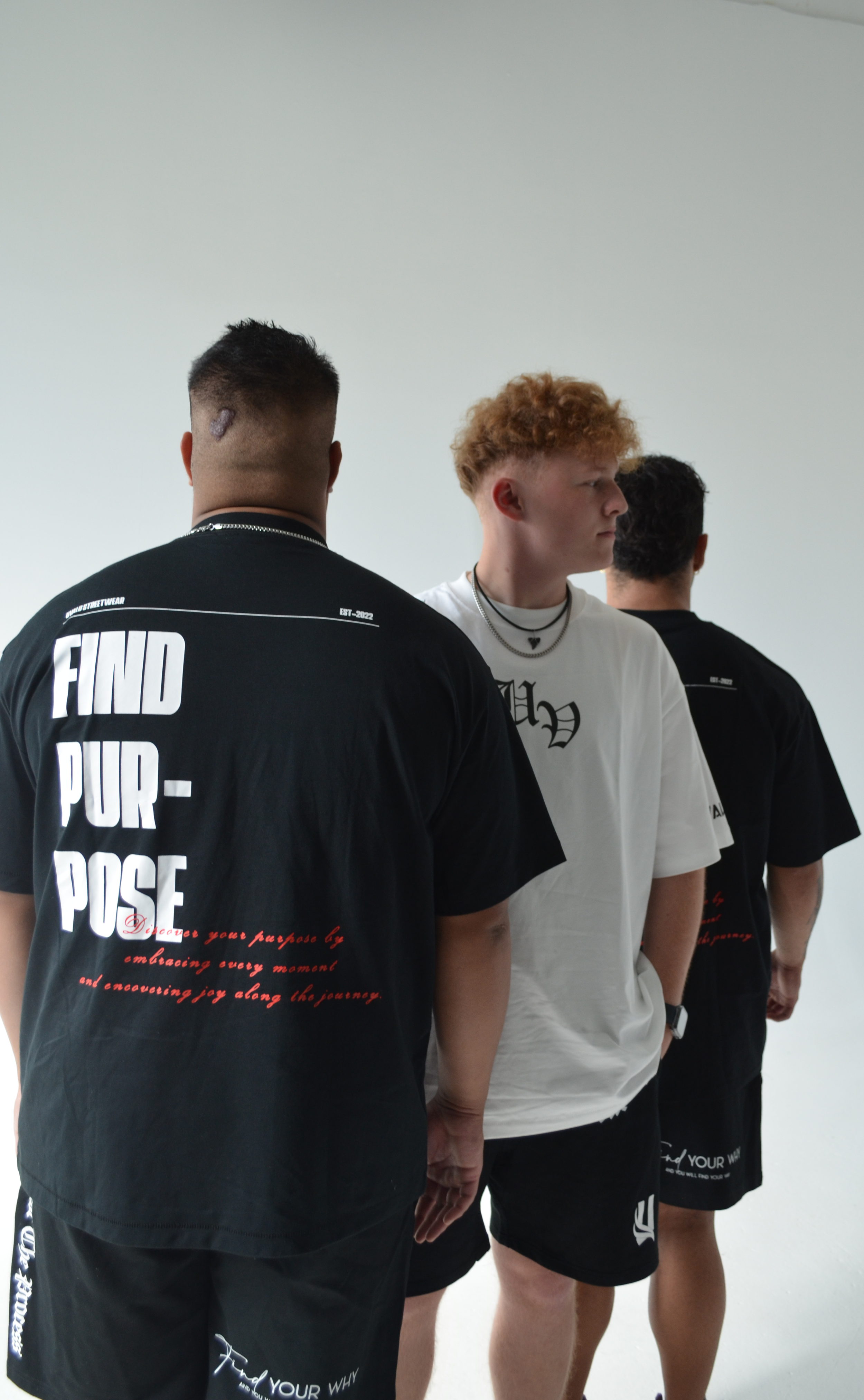 Three young men standing in a row facing away from the camera, wearing casual black and white clothing, in front of a plain white background.