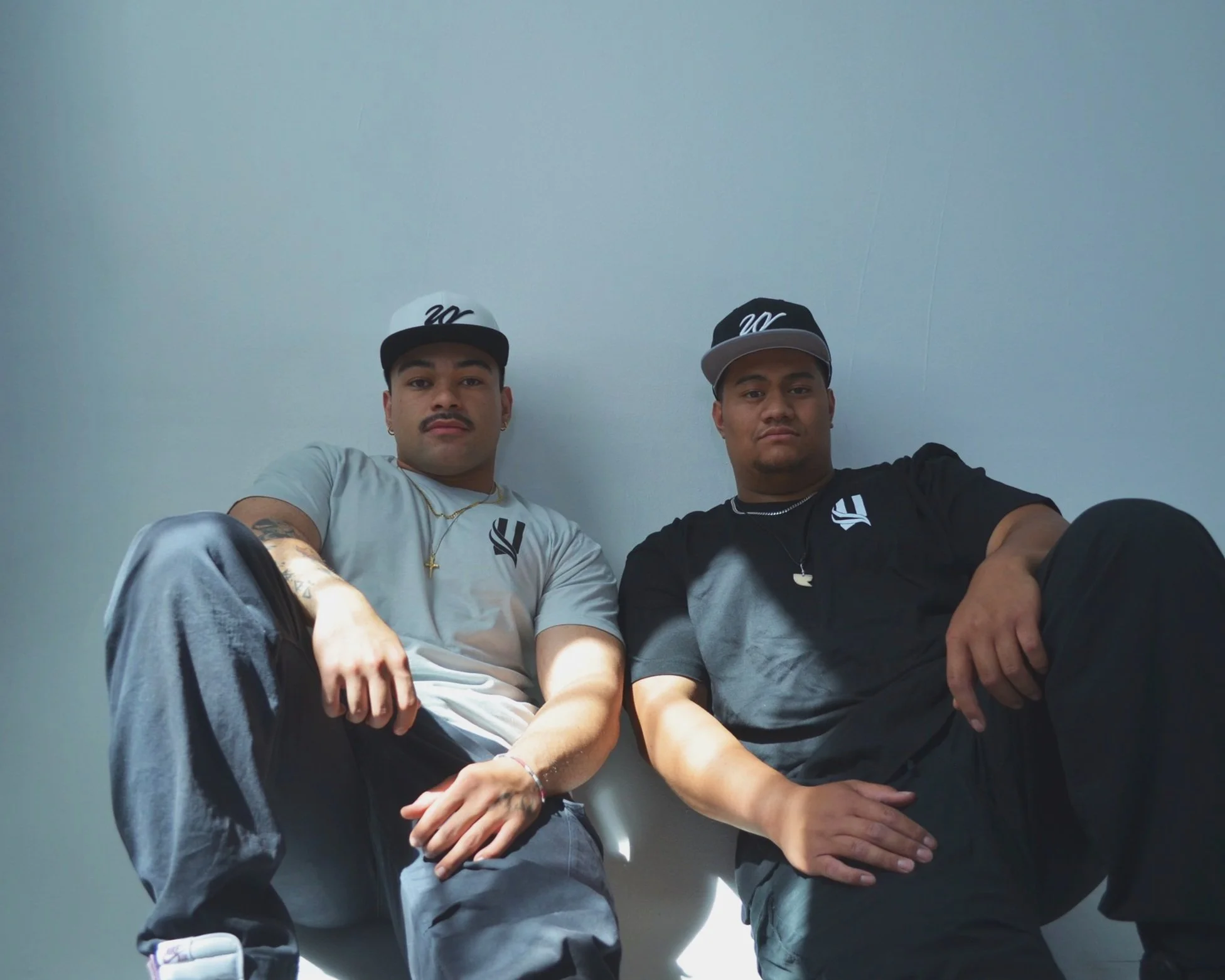 Two young men sitting against a light-colored wall, wearing casual clothing and black caps with a white logo, looking at the camera.