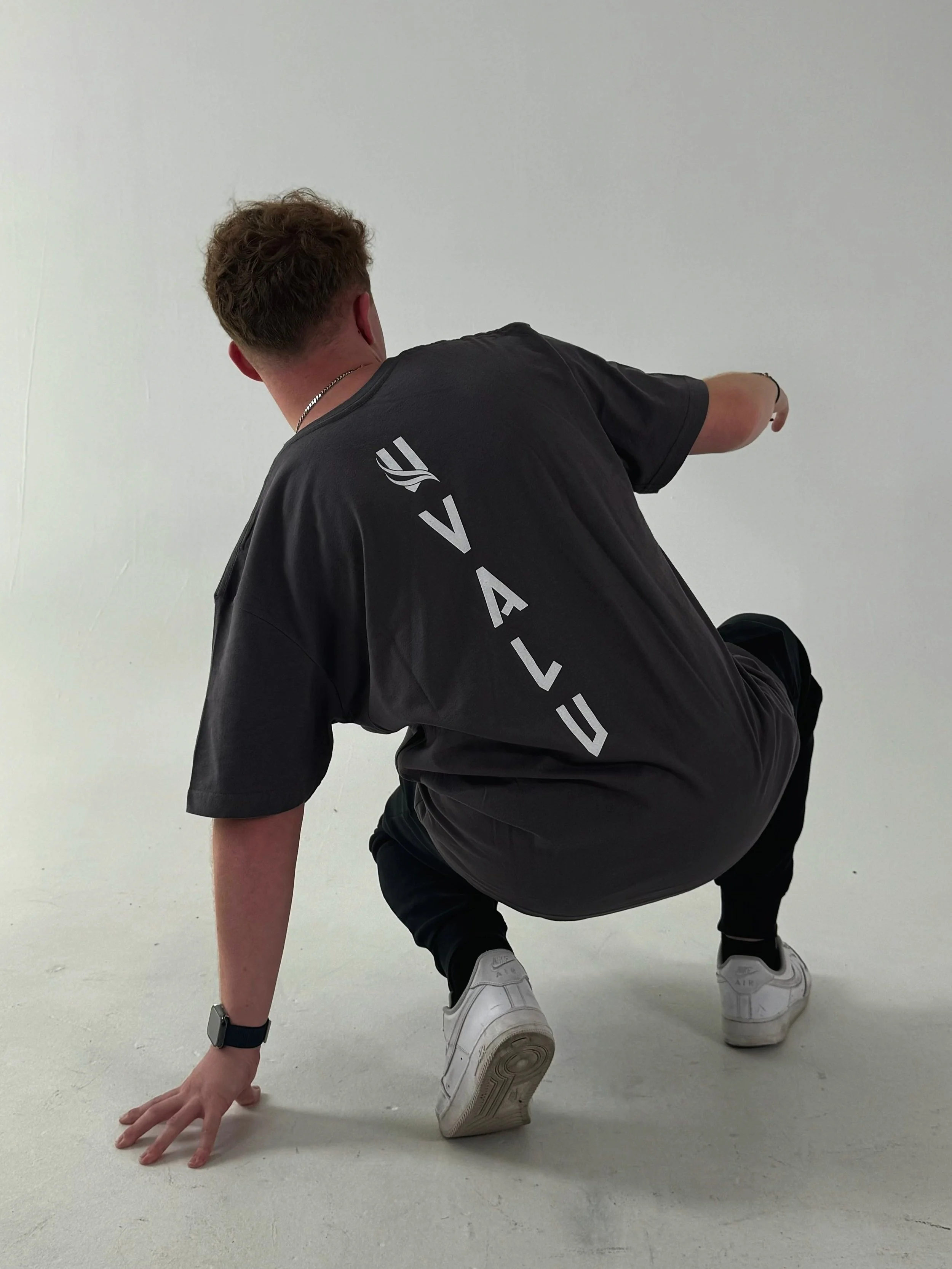 A person in a black Valu t-shirt, black pants, and white sneakers, in a crouching pose on a plain white background.