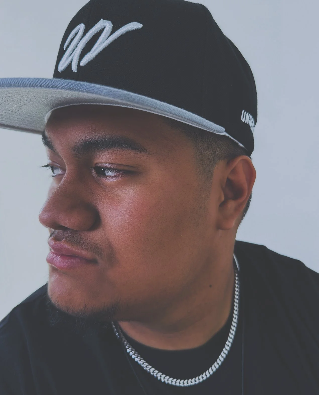 Close-up of a young man wearing a black baseball cap with a gray brim and white embroidery, a silver chain necklace, and a black shirt.