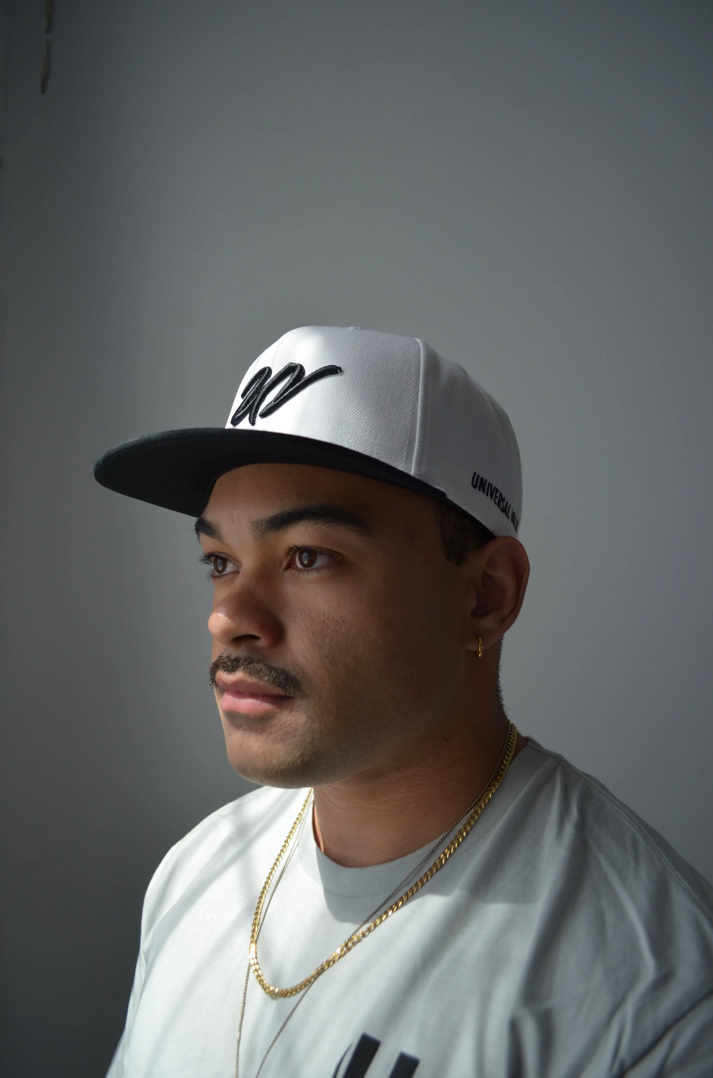 Portrait of a young man wearing a white cap with black and white lettering, gold chain necklaces, a white t-shirt, and a small gold hoop earring, looking to the left against a plain gray background.