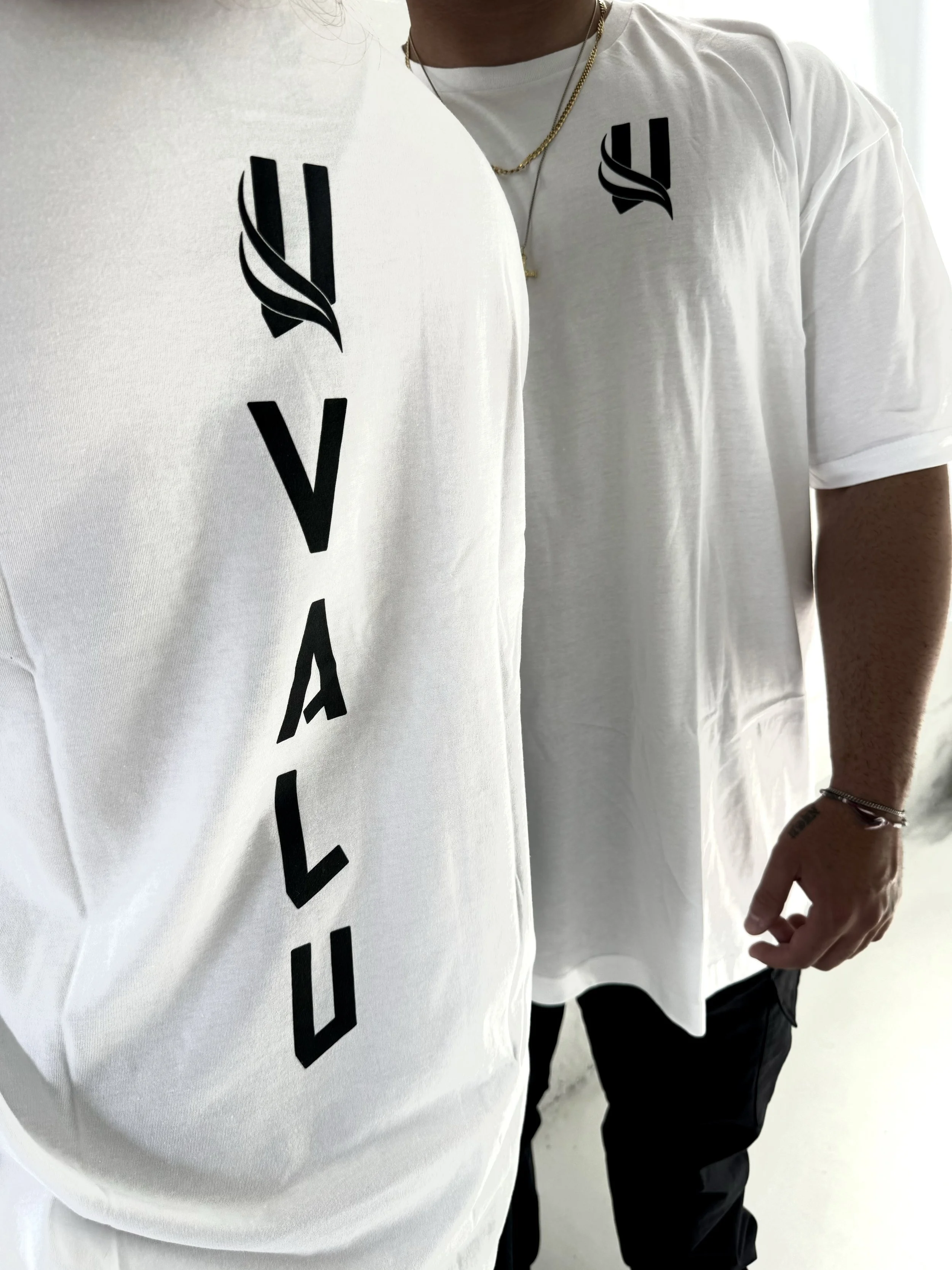 Person wearing a white T-shirt with the word 'VITAL' printed vertically and a logo on the chest, along with black pants and layered gold necklaces.