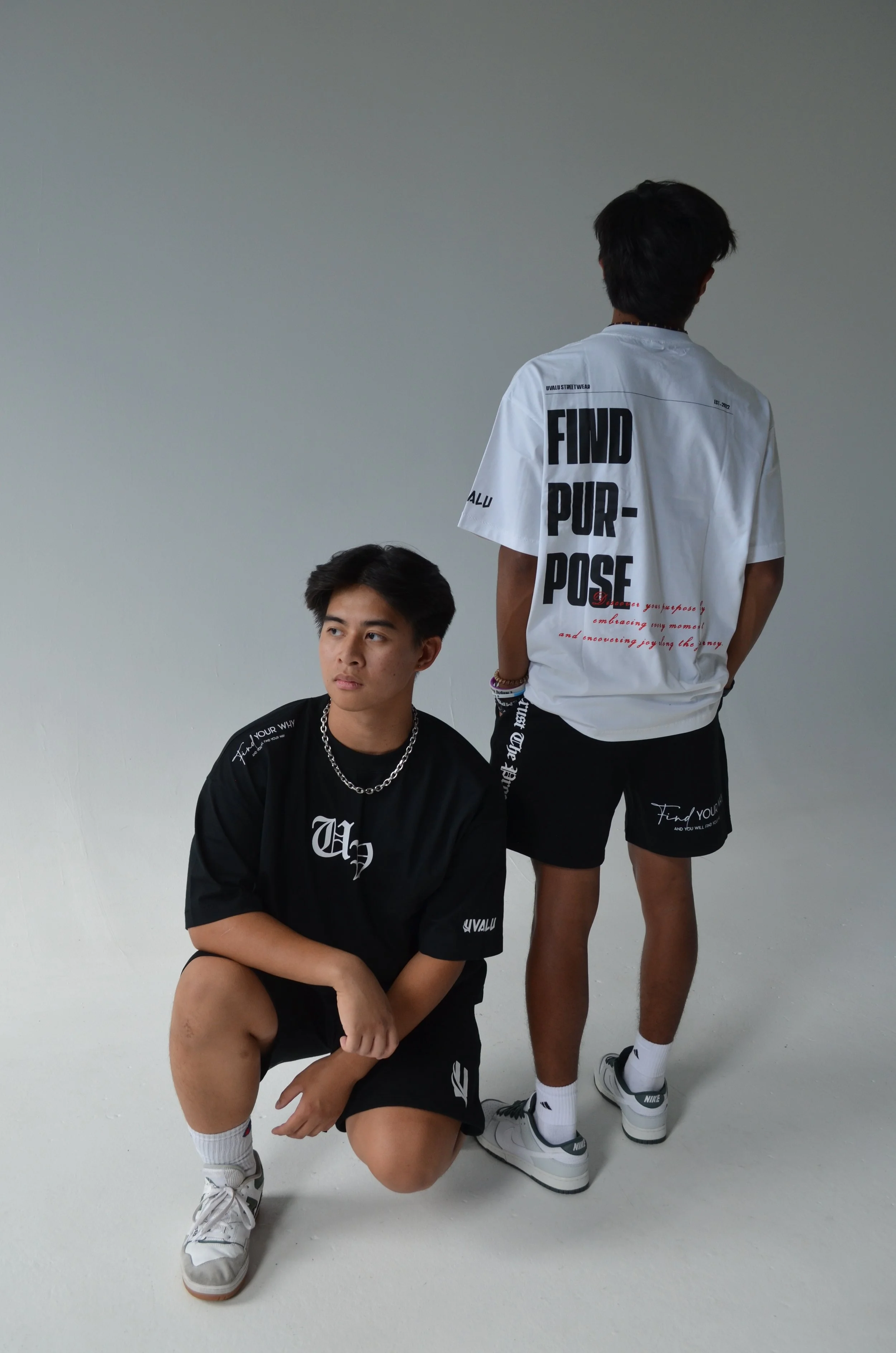 Two young men in casual streetwear pose against a plain white background. One is crouching with a black outfit, and the other is standing with his back to the camera, wearing a white t-shirt and black shorts.