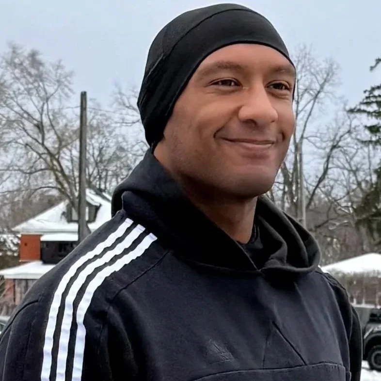Smiling man in black sportswear with white stripes on the shoulder, wearing a black head covering, outdoors in winter with snow and trees in the background.