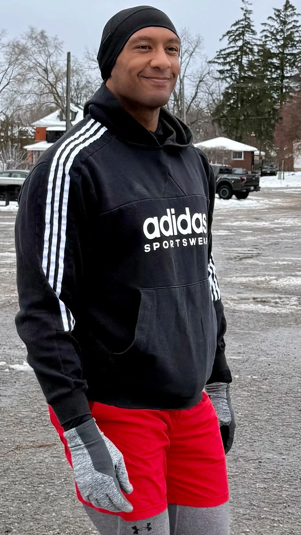 A man dressed in black Adidas sportswear with a black headband and red shorts, standing outdoors in a snowy parking lot during winter.