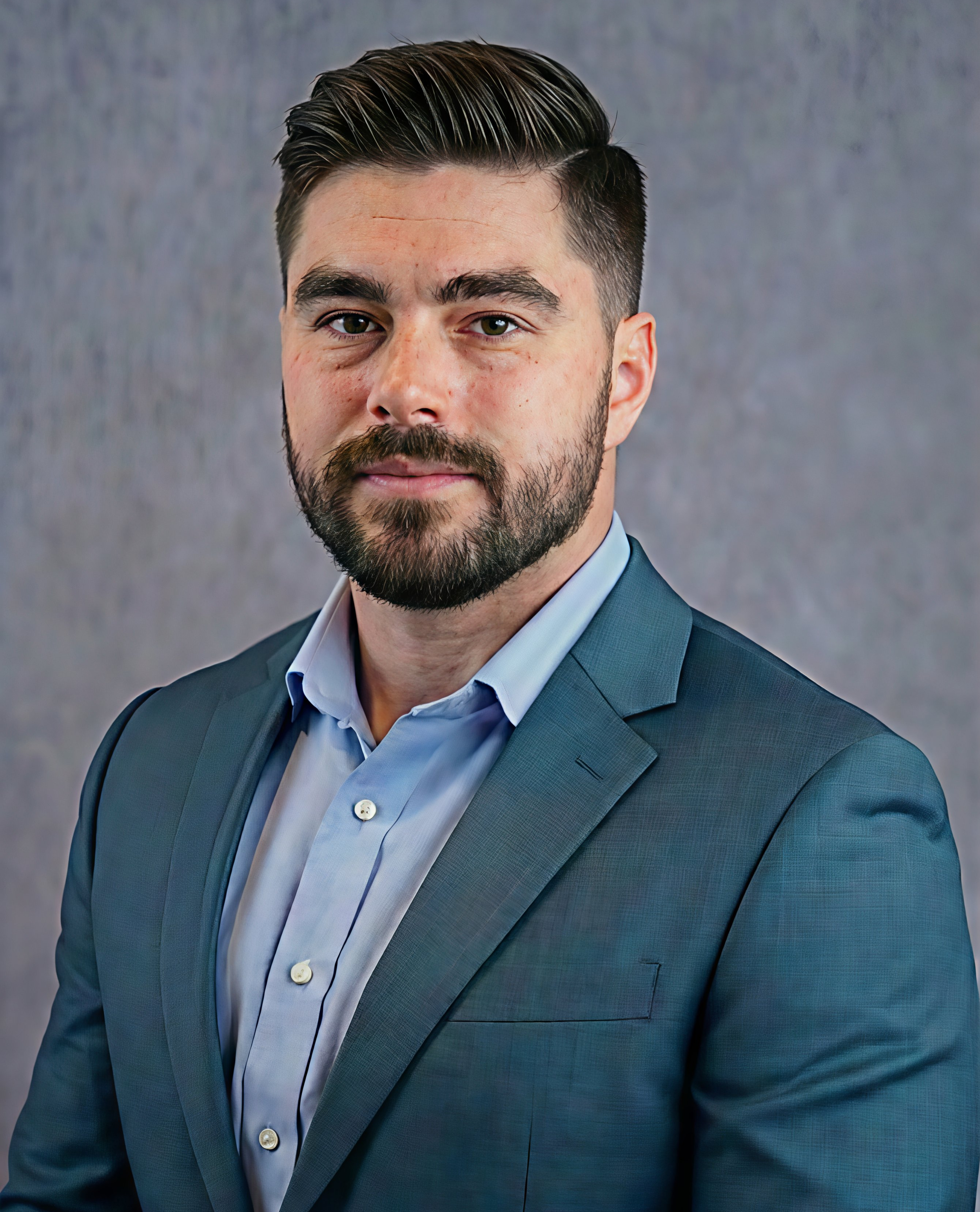 Professional man wearing a suit and light blue shirt, looking at camera with a serious expression, against a plain gray background.