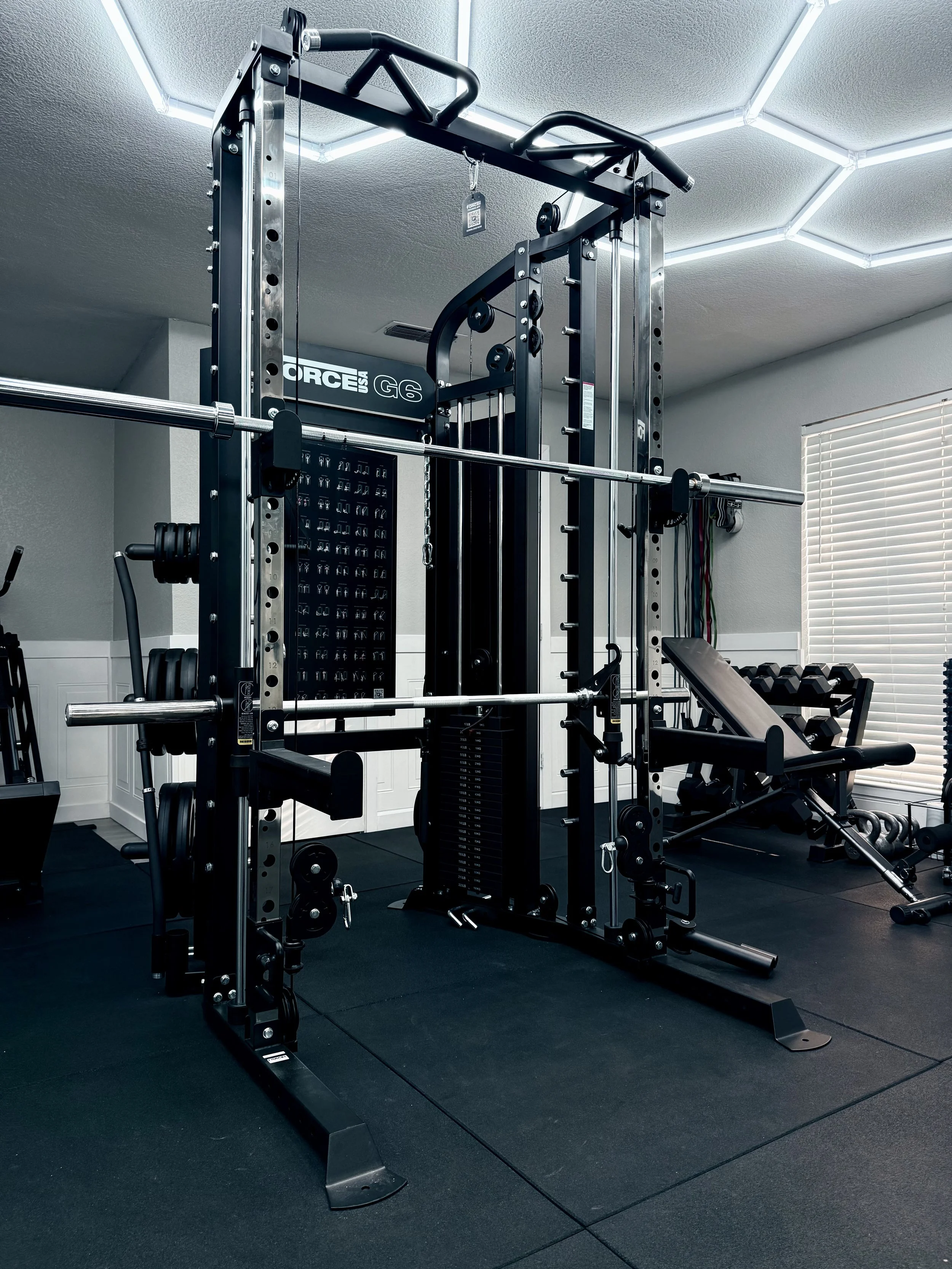 Home gym with a multi-functional exercise machine, weight plates, and a weight bench, located in a well-lit room with white walls and blinds.