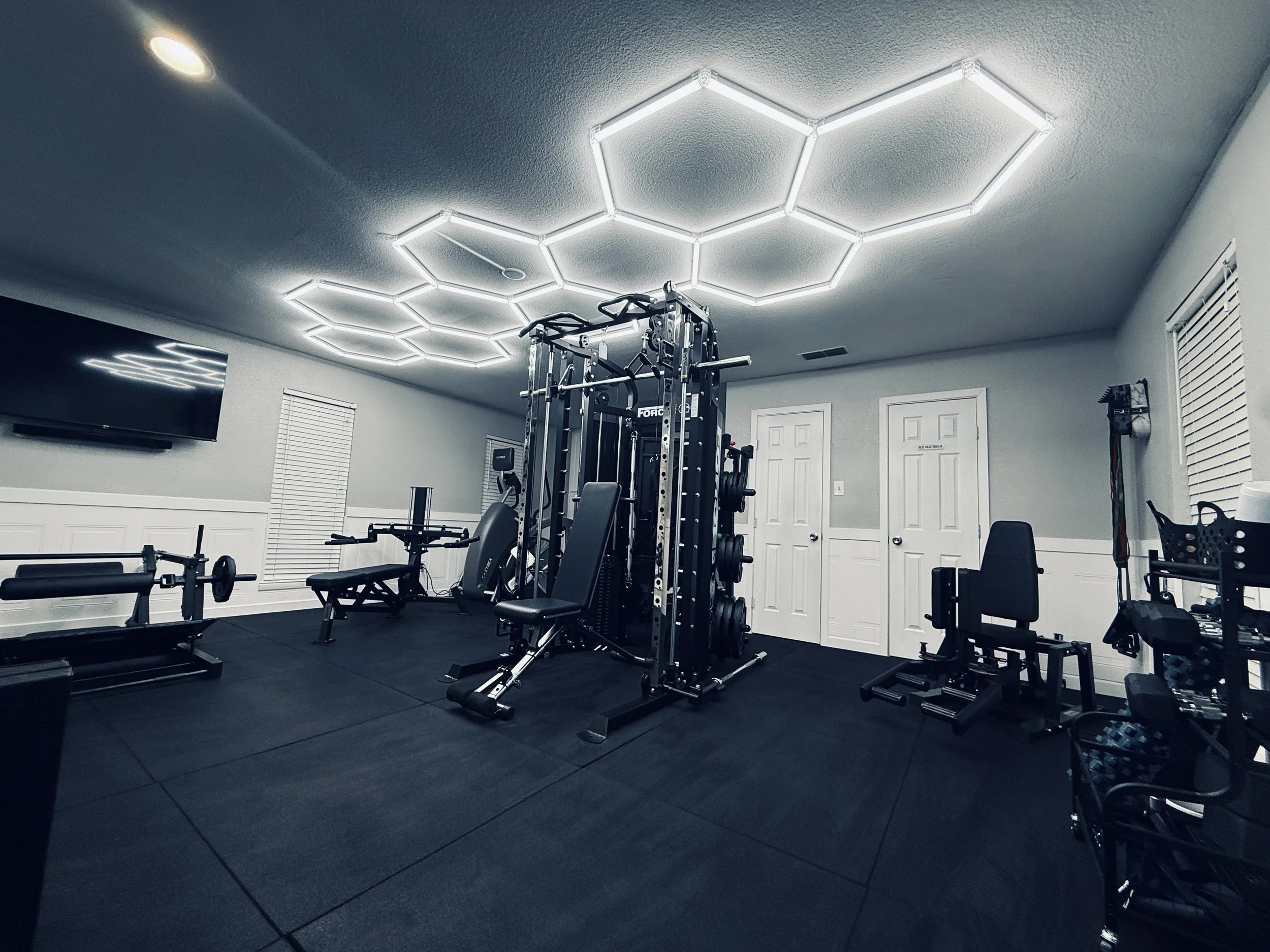 Home gym with various workout equipment including a weightlifting rack, benches, a TV on the wall, and hexagon-shaped LED ceiling lights.