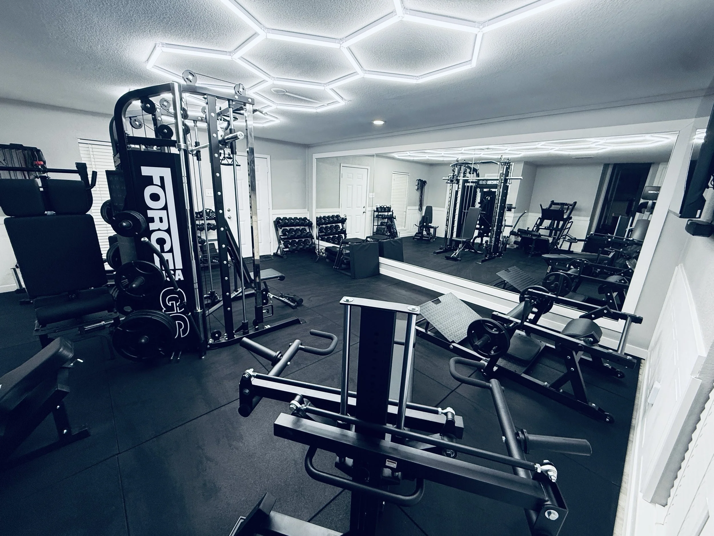 A home gym with fitness equipment, including weight benches, dumbbell racks, and a mirrored wall. The ceiling has modern hexagonal light fixtures and the room has gray walls and black flooring.