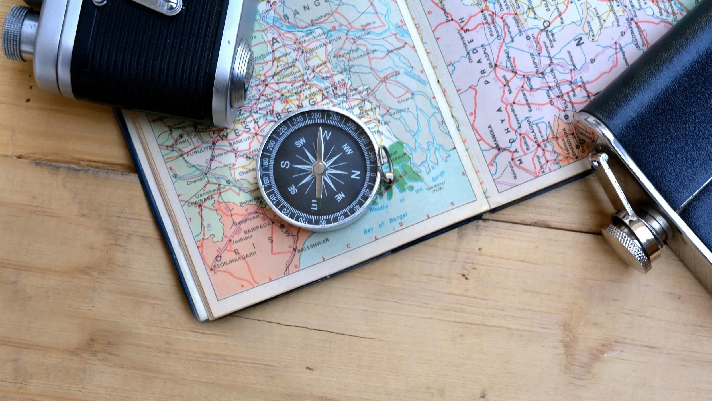 Travel items on a wooden surface, including a map, compass, binoculars, and a camera.