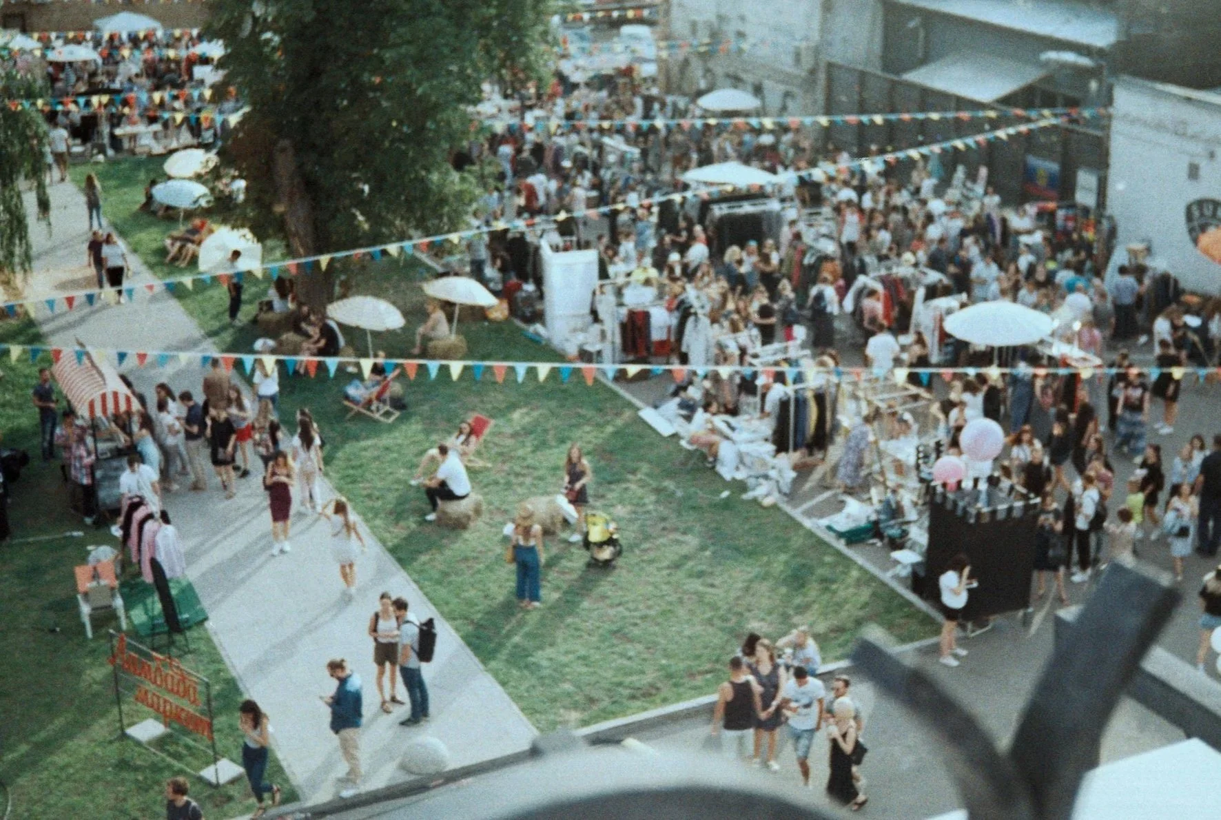 Bird's eye view of a crowded outdoor market with street vendors, shoppers, and decorative buntings. There are tables with clothes, umbrellas, and people walking around.