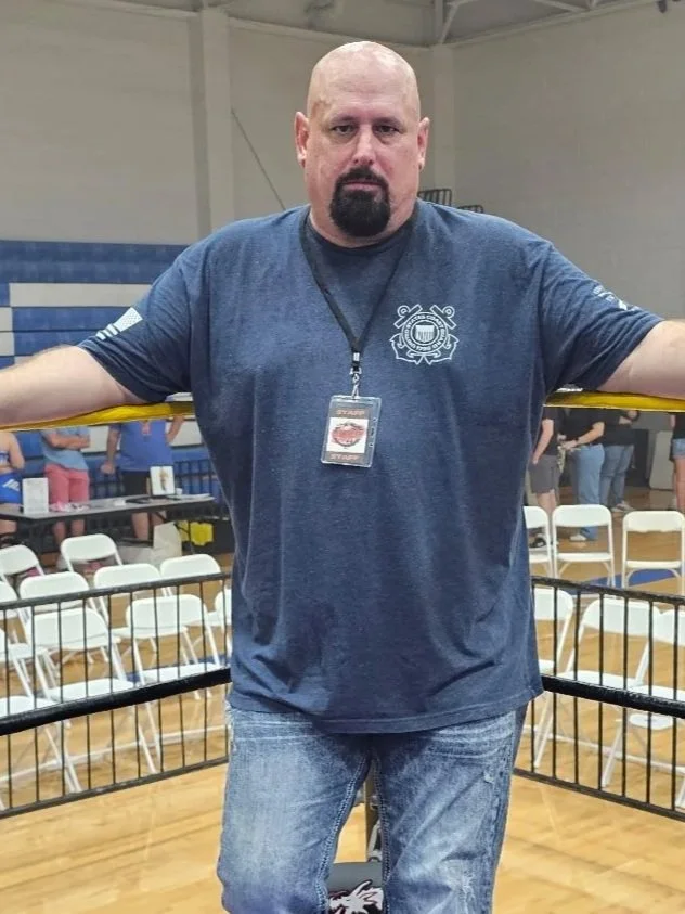 A bald man with a goatee, wearing a navy t-shirt with a badge around his neck, standing inside a boxing ring with white chairs and tables in the background.