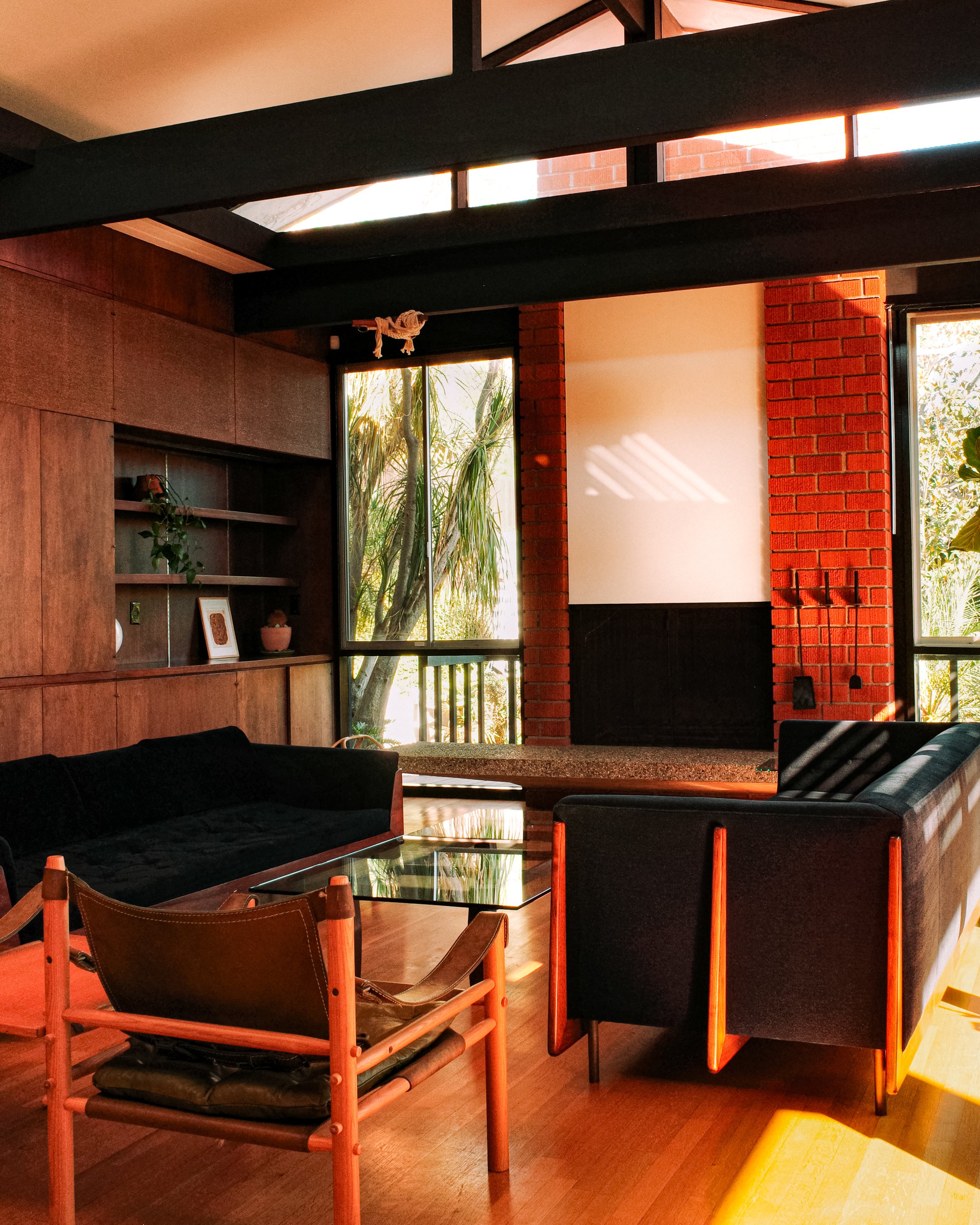 A cozy living room with wooden flooring, black sofas, a glass coffee table, and a built-in wooden bookshelf with decorative items. There are large windows with sunlight streaming in, and a brick wall accent with a small fireplace. A yellow chair is partially visible in the foreground.