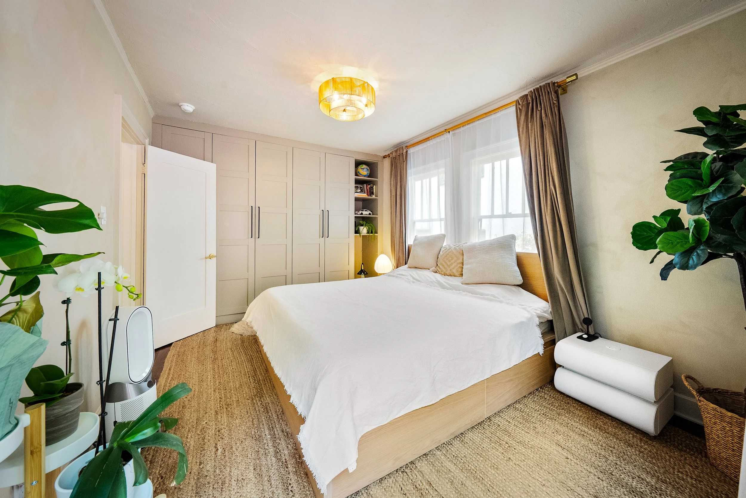 A cozy bedroom with a large bed, white bedding, and two beige pillows. There's a white wardrobe, a bookshelf with books and a globe, and several potted plants. Large windows with sheer curtains and beige drapes let in natural light. A modern ceiling light fixture is on the ceiling.