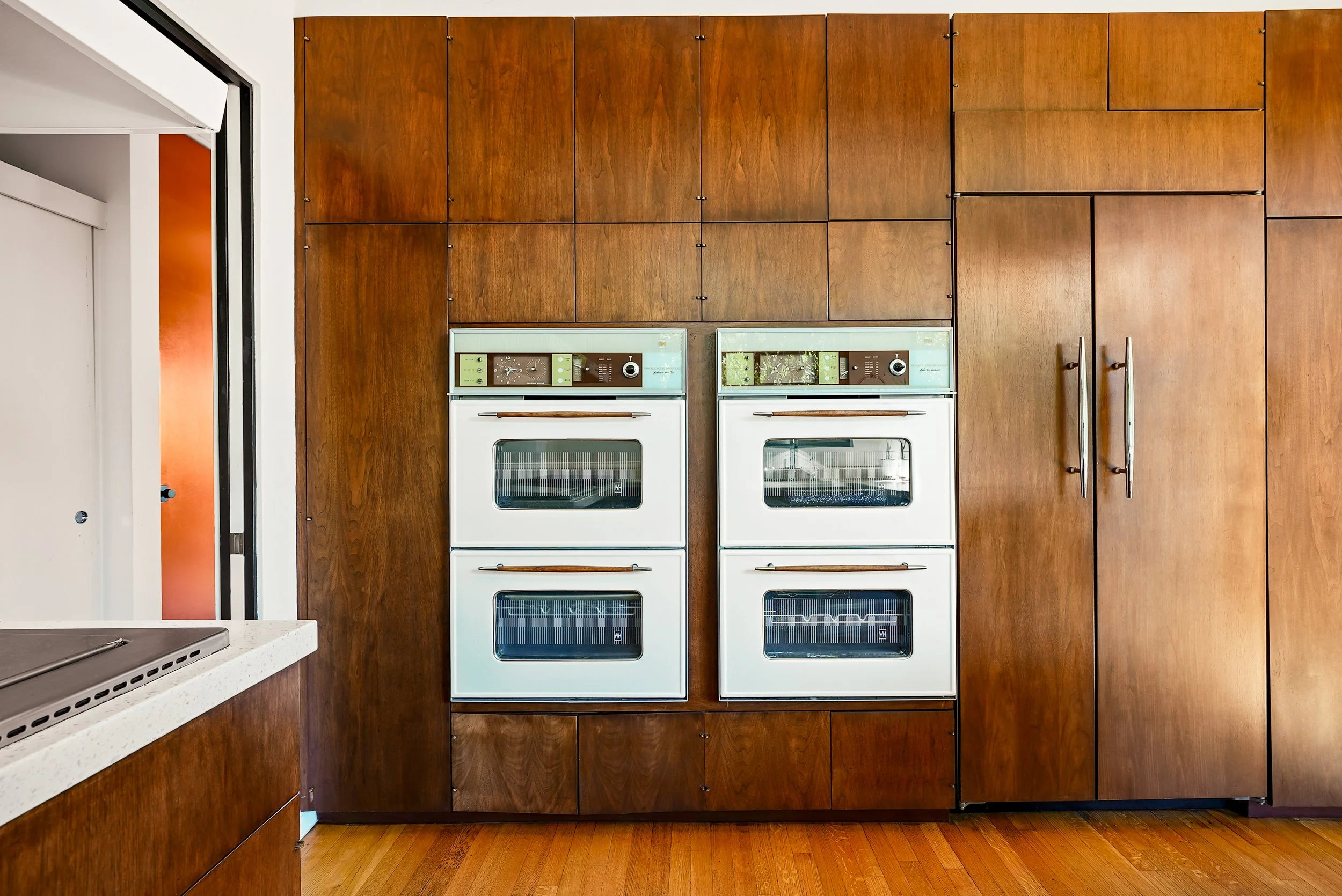Kitchen with wooden wall cabinets and a matching floor. Two vintage white ovens are built into the wall, next to a purple refrigerator. Part of a white countertop and sink are visible in the foreground.