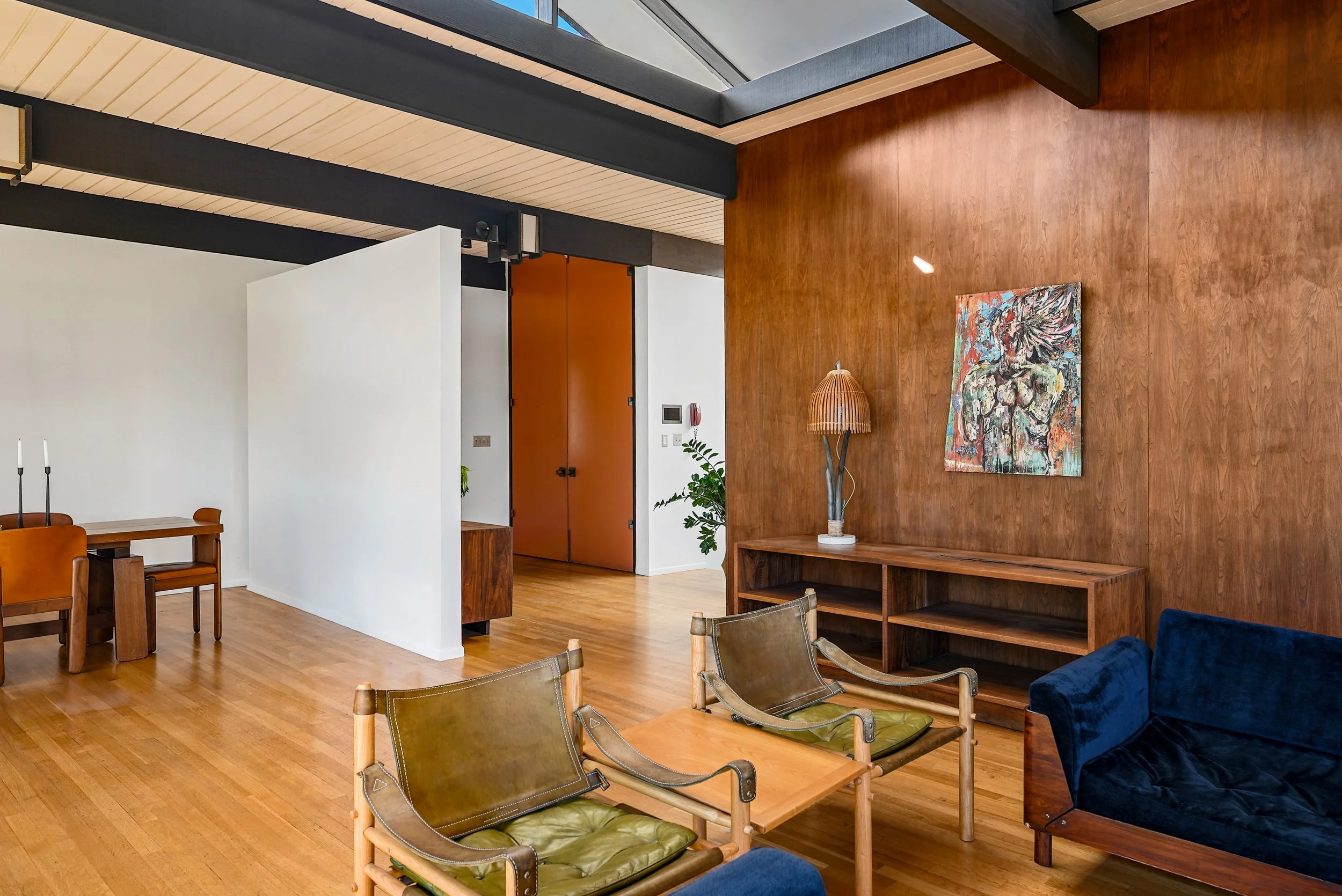 Modern living room with wooden floor, wooden wall, and mid-century furniture. Features a blue velvet velvet armchair, wooden chairs, a wooden shelf, an abstract painting, a floor lamp, and some houseplants.