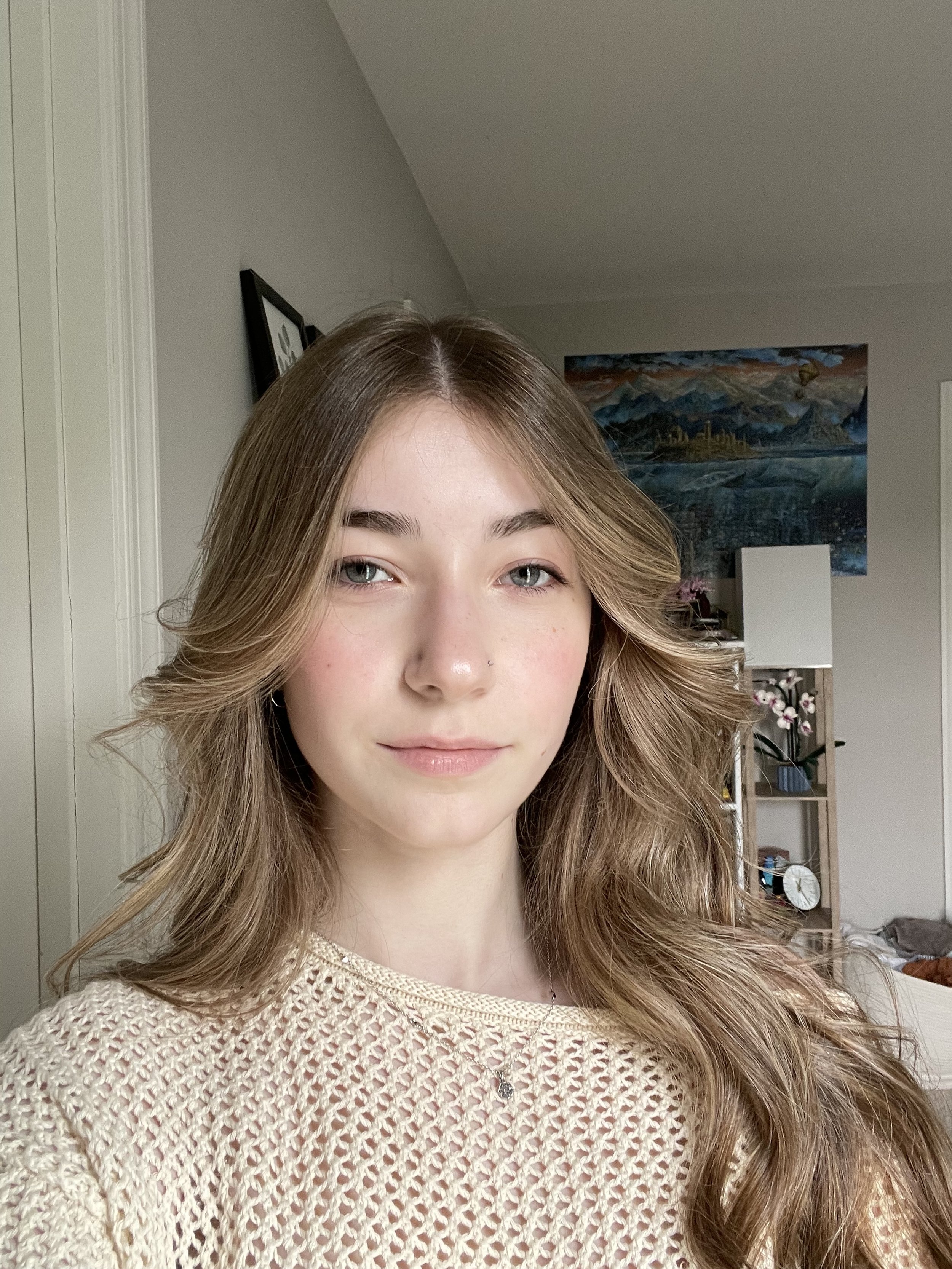 A young woman with long, wavy light brown hair and blue eyes, wearing a beige crochet top, standing indoors in a well-lit room with artwork and plants in the background.