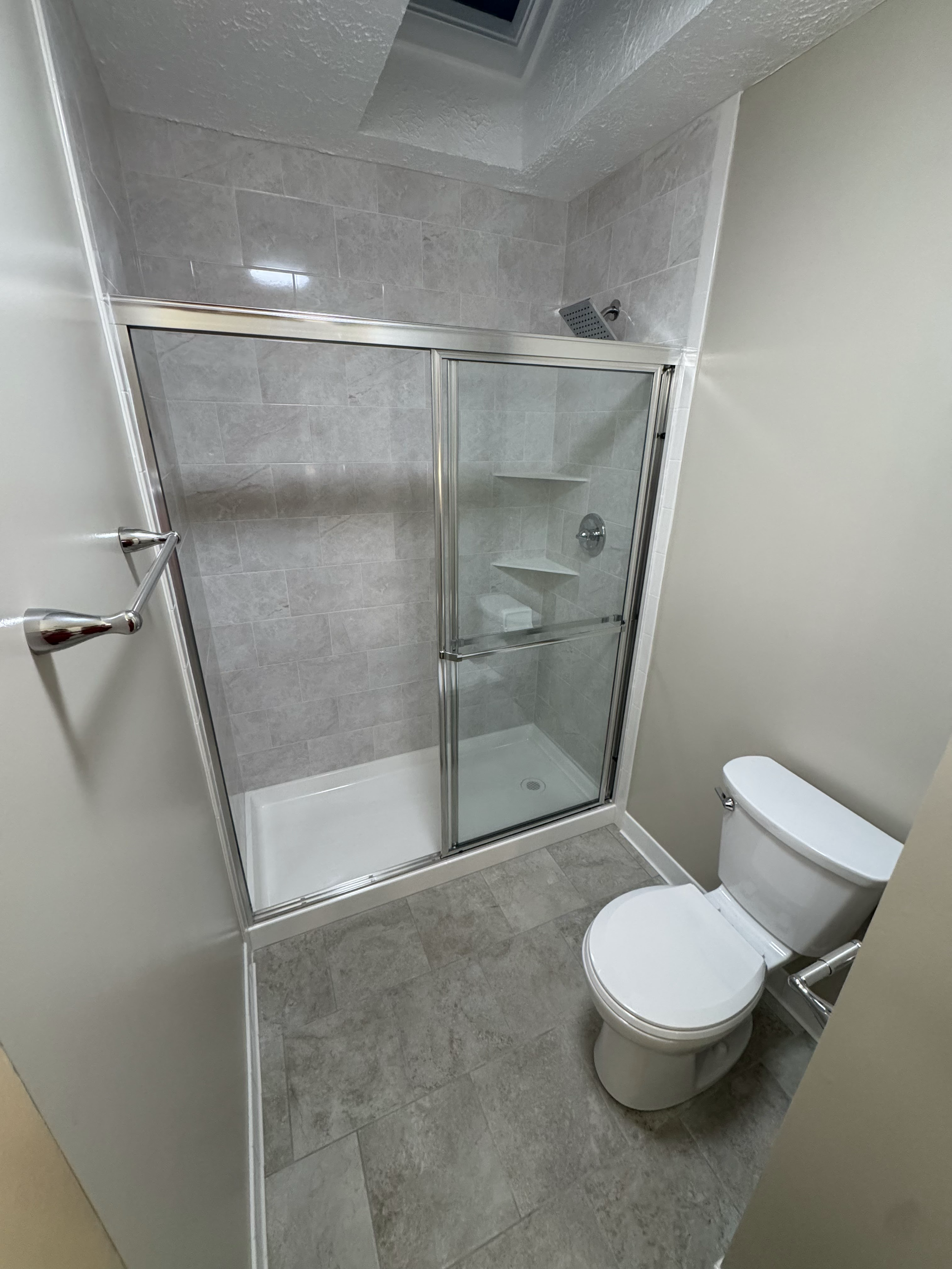 A small bathroom featuring a glass shower enclosure with tiled walls, a showerhead, a drain, and built-in shelves, next to a white toilet, with beige walls and gray tile flooring.