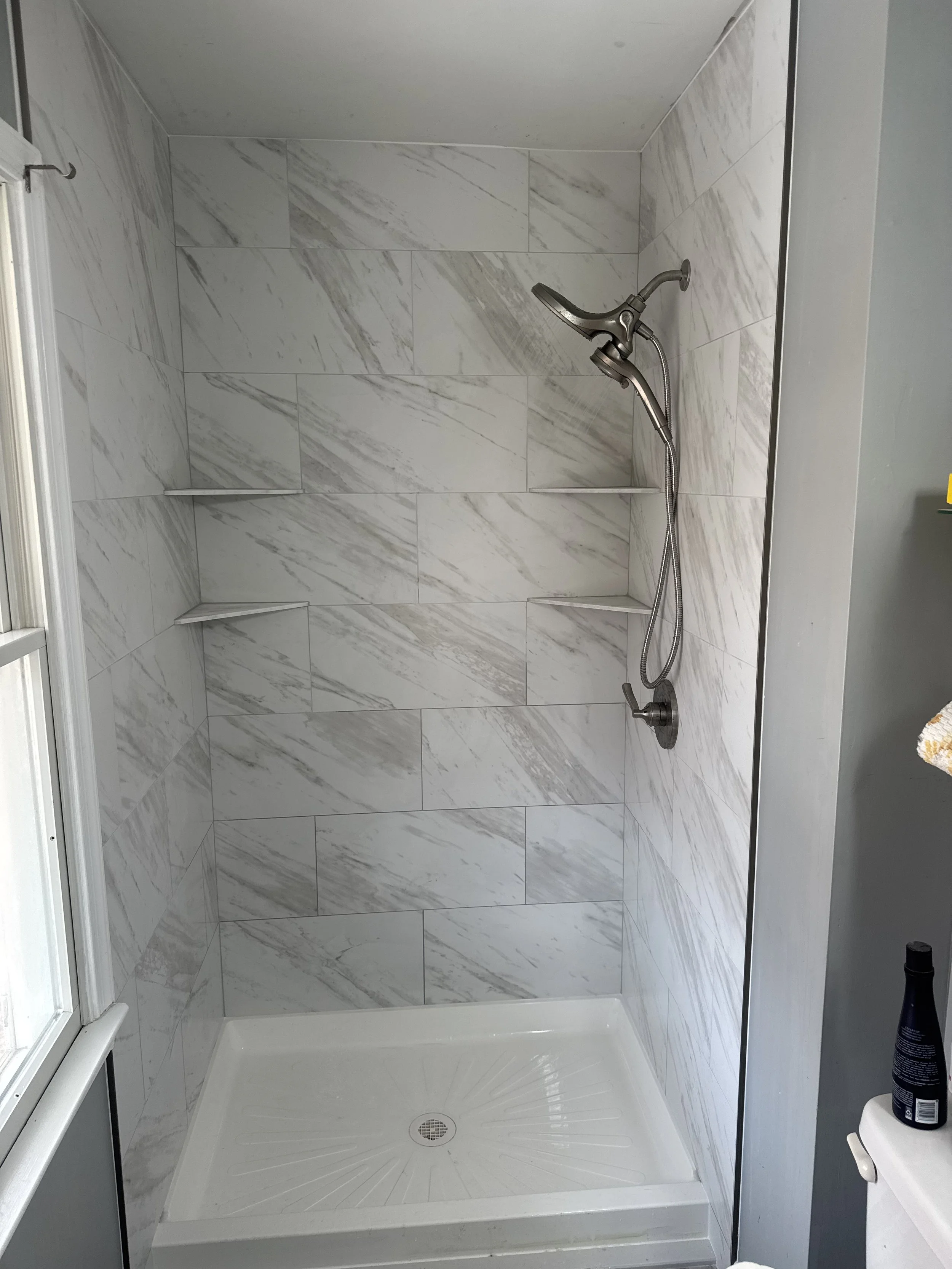 Marble-tiled shower with a handheld showerhead, built-in corner shelves, and a white shower base with a drainage grate.