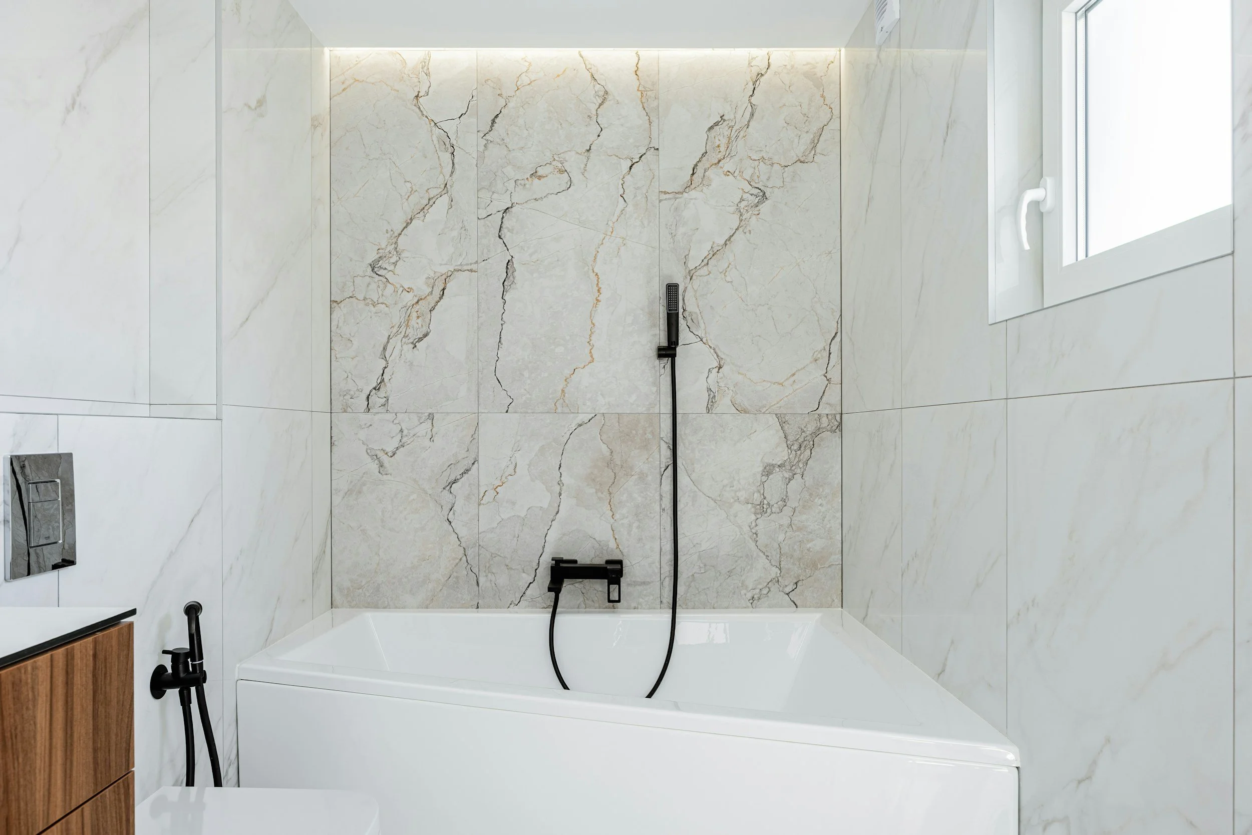 Modern bathroom with marble tiles on the wall, a white bathtub, a black handheld shower, a small window, and minimalist fixtures.