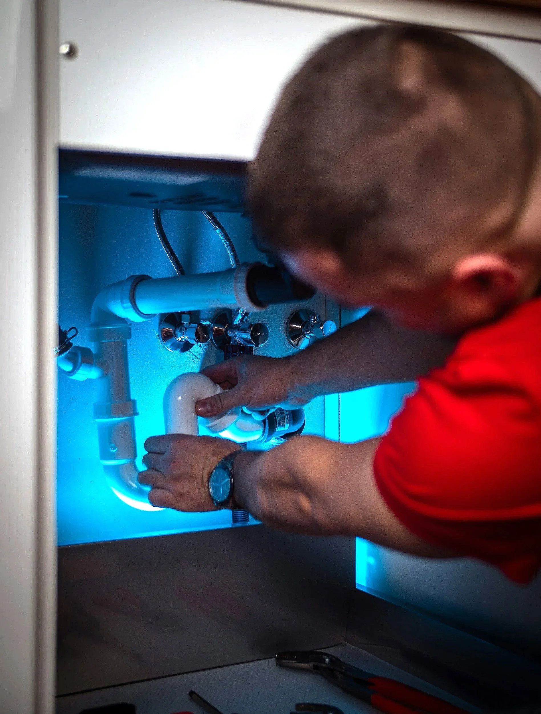 Hands working under a kitchen sink illuminated by blue light, with plumbing pipes and tools visible.