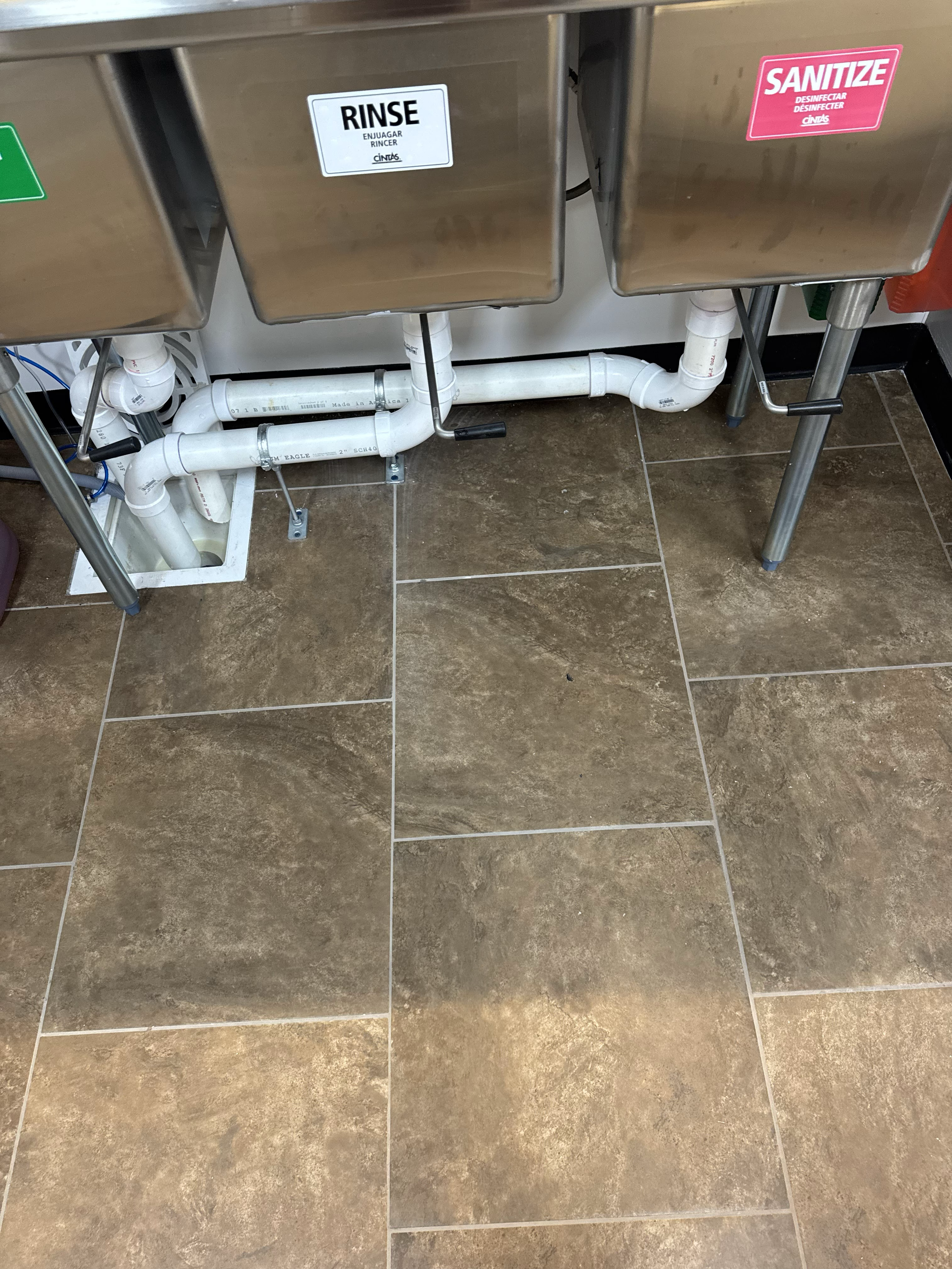 View of plumbing pipes beneath stainless steel sinks with labels 'RINSE' and 'SANITIZE'; tiled floor in a commercial or industrial setting.