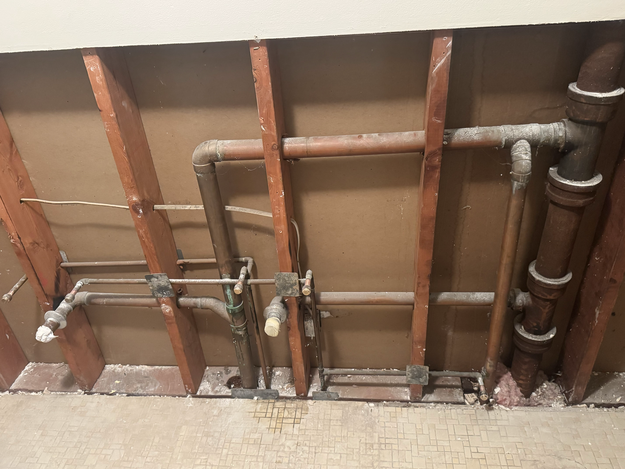 Inside view of a bathroom wall showing exposed plumbing pipes and wooden framing, with some drywall removed.