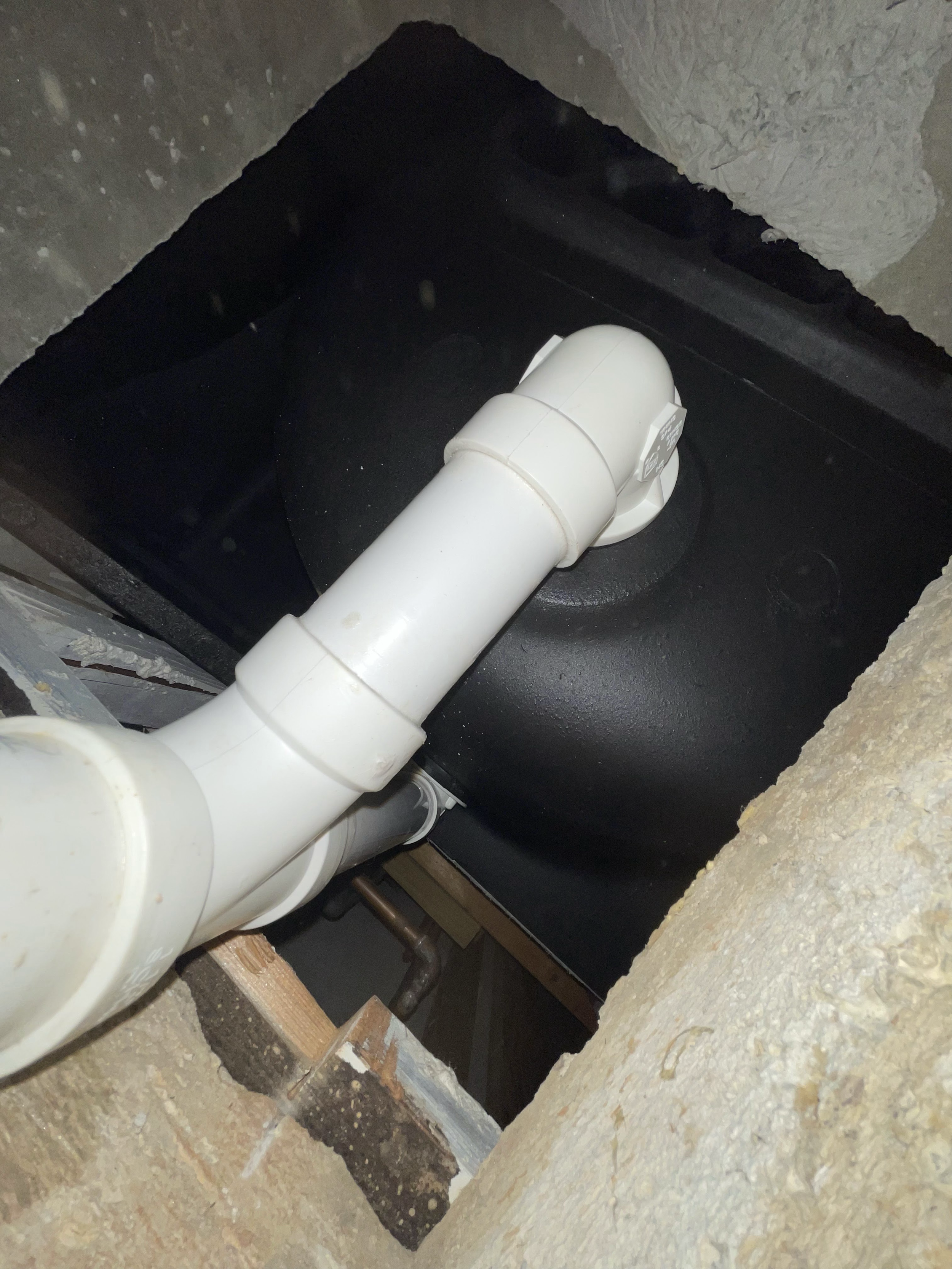 A close-up view of plumbing pipes installed within a wall opening, showing a white PVC pipe connected to a black tank or component.