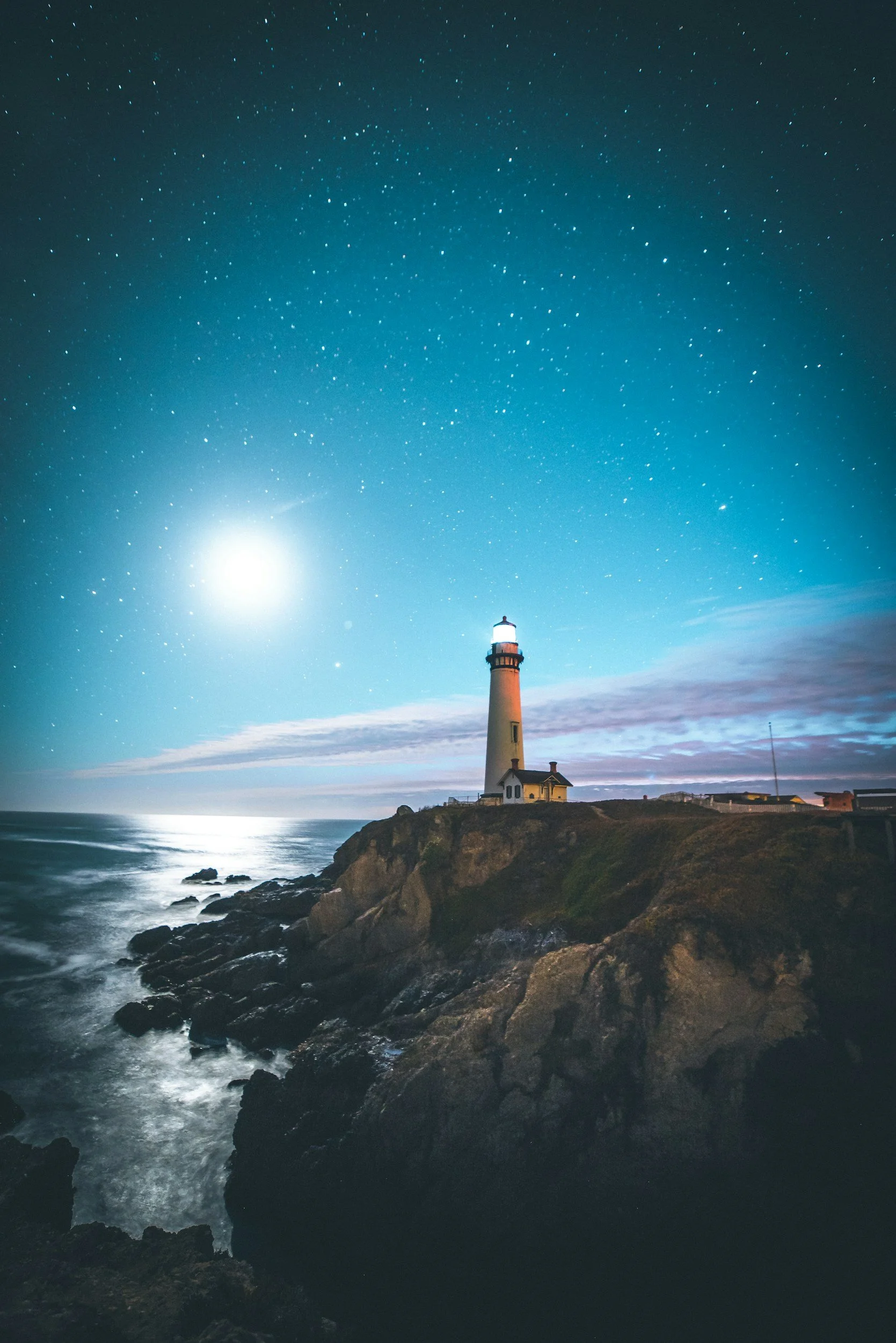 Nighttime scene of a lighthouse on a rocky coast with a bright moon and starry sky reflected on the water.