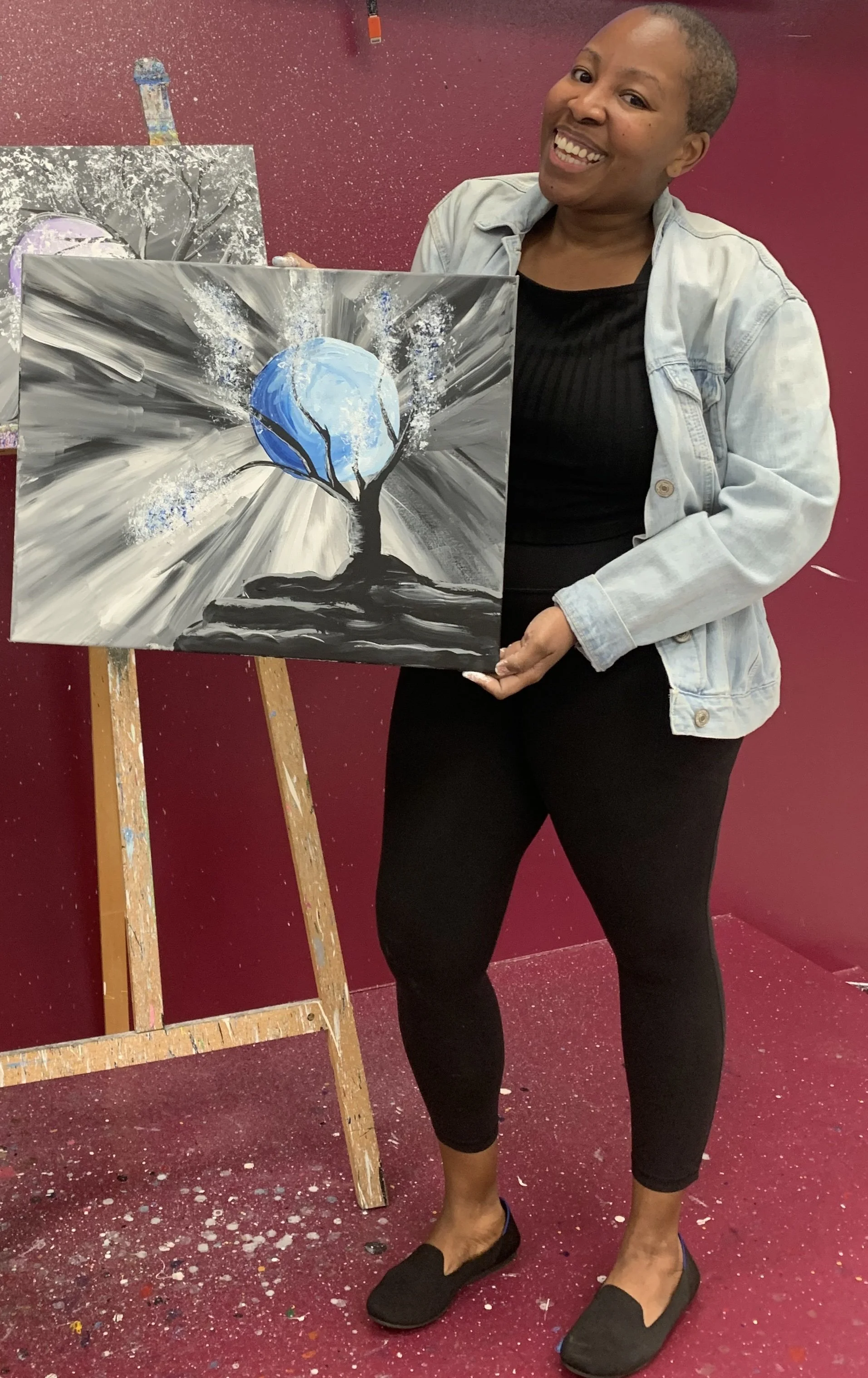 Sunny, a Black woman holding an abstract painting of a tree and moon on a canvas at an art studio. She is smiling and standing next to an easel with red walls and paint splatters on the floor.