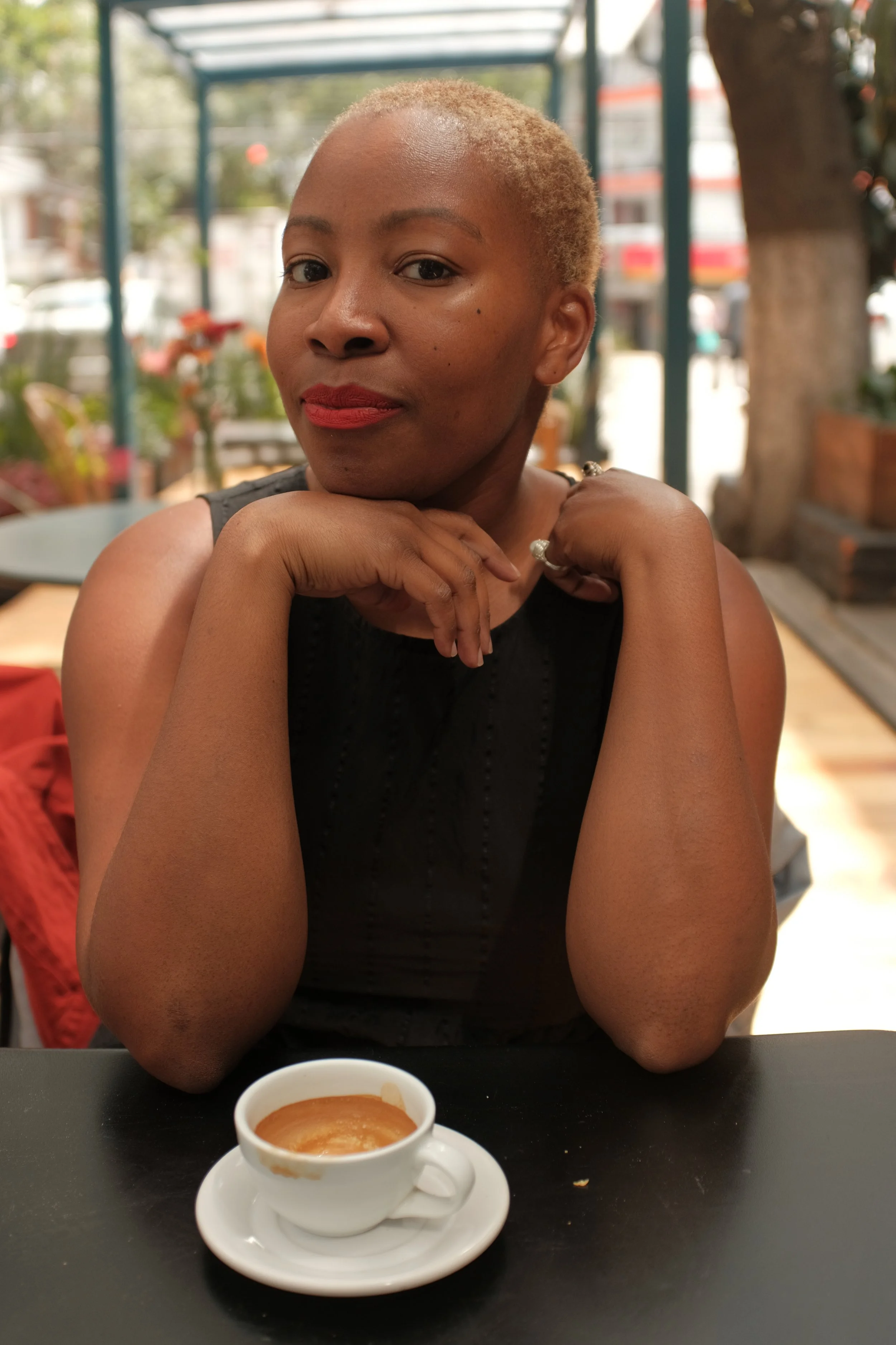 Sunny, a Black woman with short blonde hair and red lipstick seated at a table with a small cup of espresso in front of her, in an outdoor café setting.
