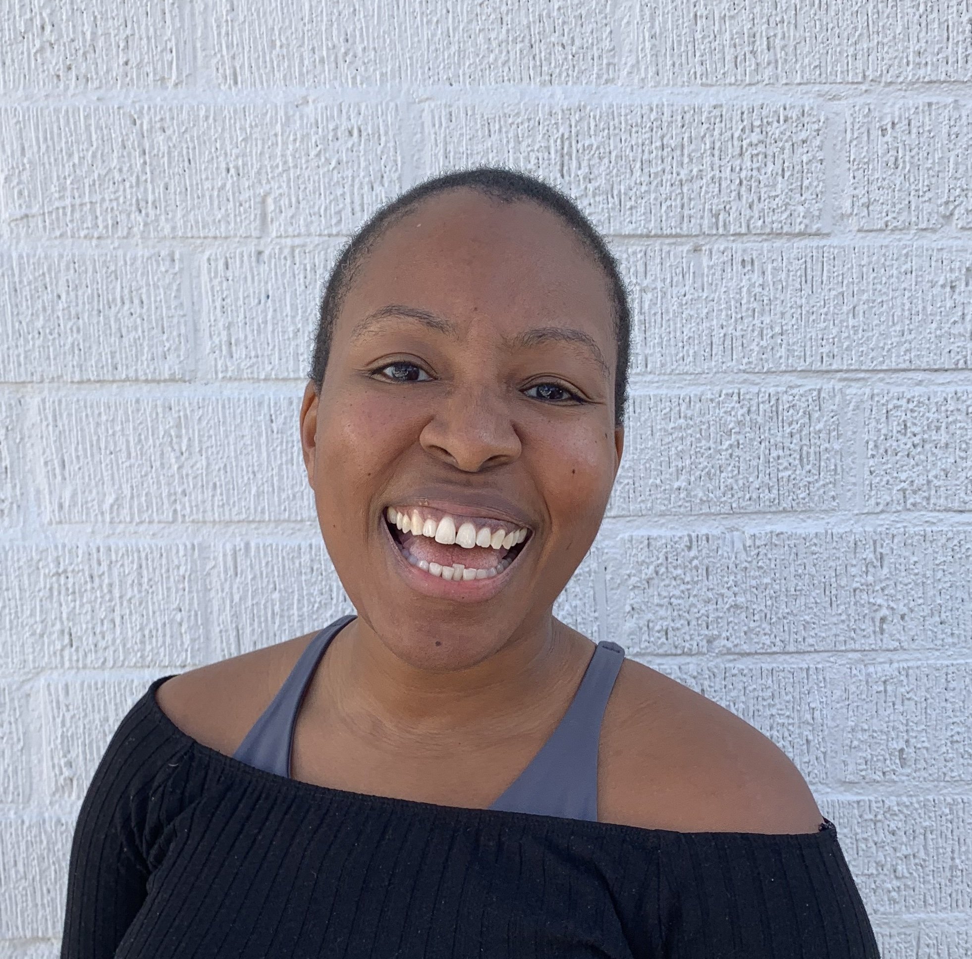 Sunny, a smiling Black woman with a shaved head, wearing a black off-the-shoulder top and a gray sports bra, standing against a white brick wall.