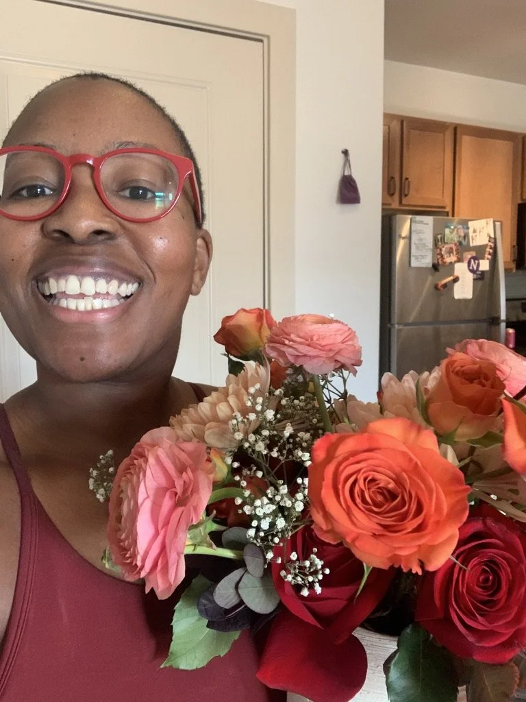 Sunny, a Black woman smiling with red glasses holding a bouquet of pink, orange, and red roses inside a home kitchen.