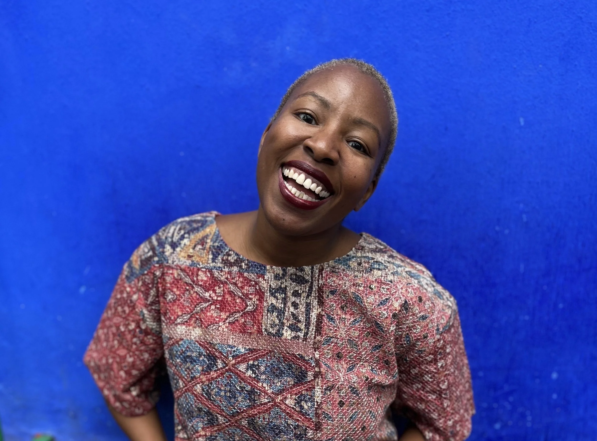Sunny, a Black woman with short hair, smiling widely showing her teeth, standing against a bright blue background, wearing a colorful patterned top.