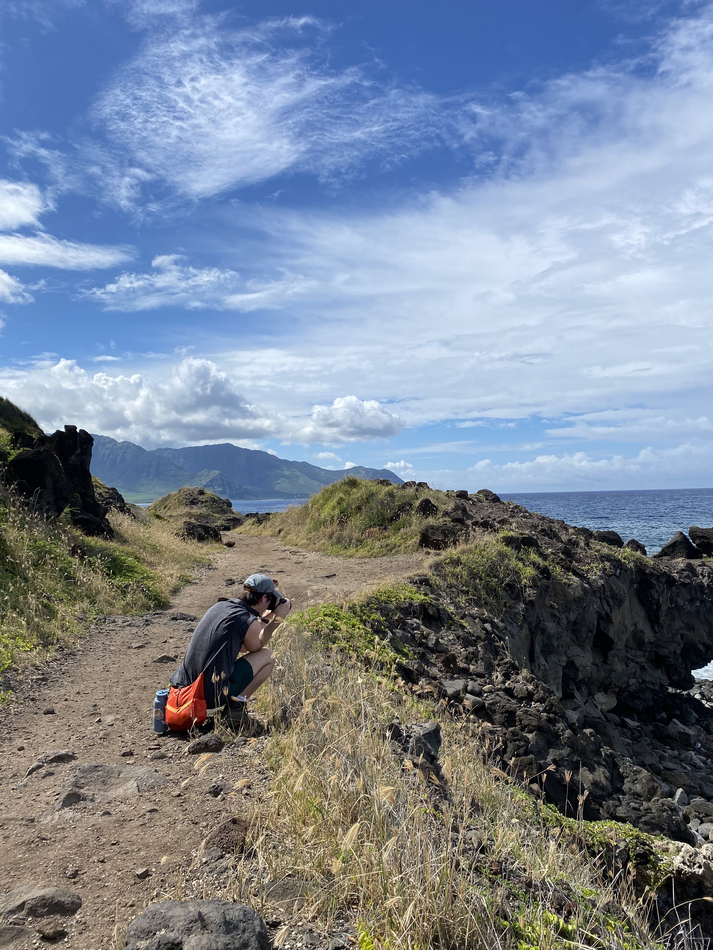 Person kneeling on a dirt trail along a rocky coastline, taking a photo of the landscape with mountains, clouds, and the ocean in the background.