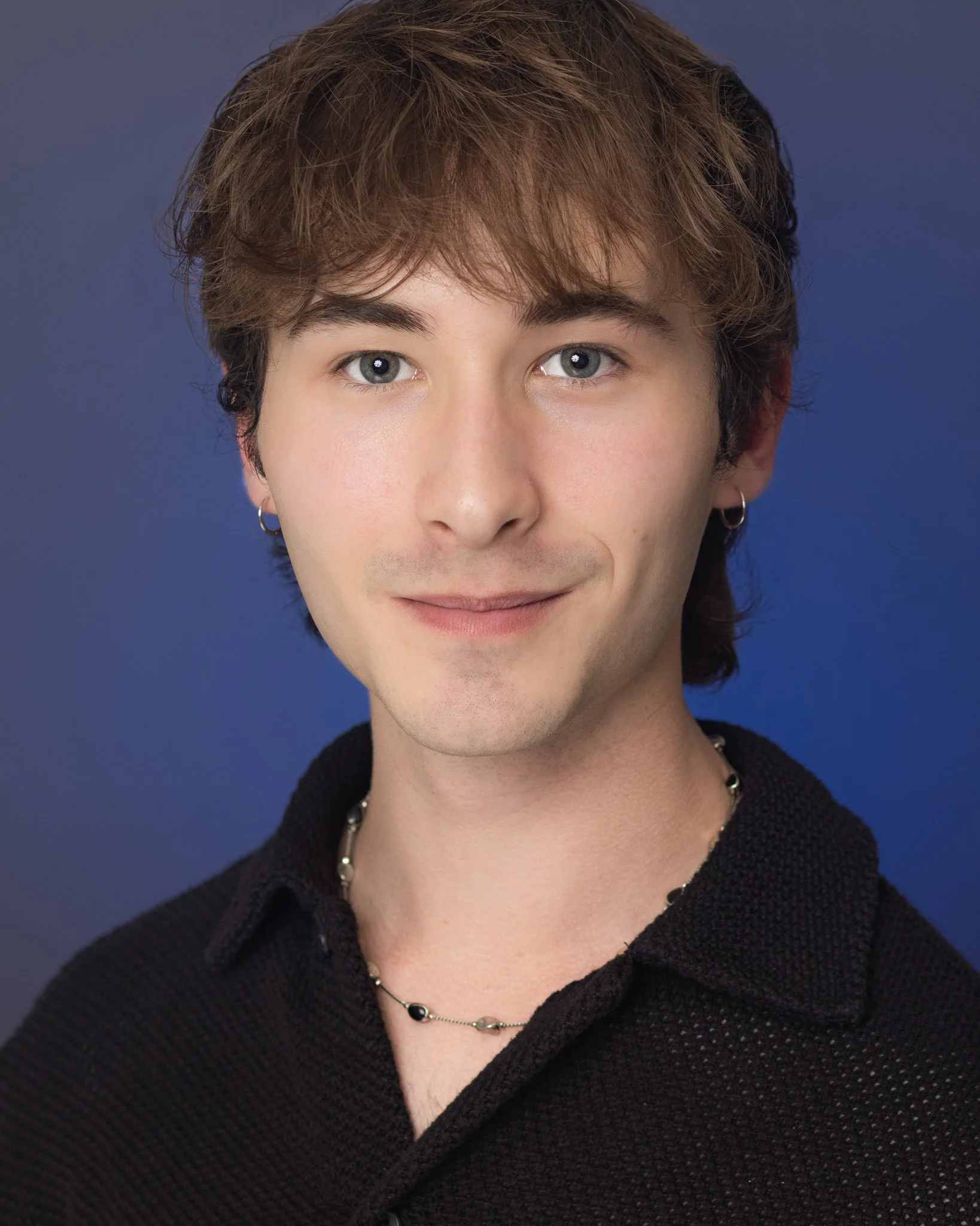 Portrait of a young man with brown hair, blue eyes, wearing earrings and a necklace, smiling against a blue background.