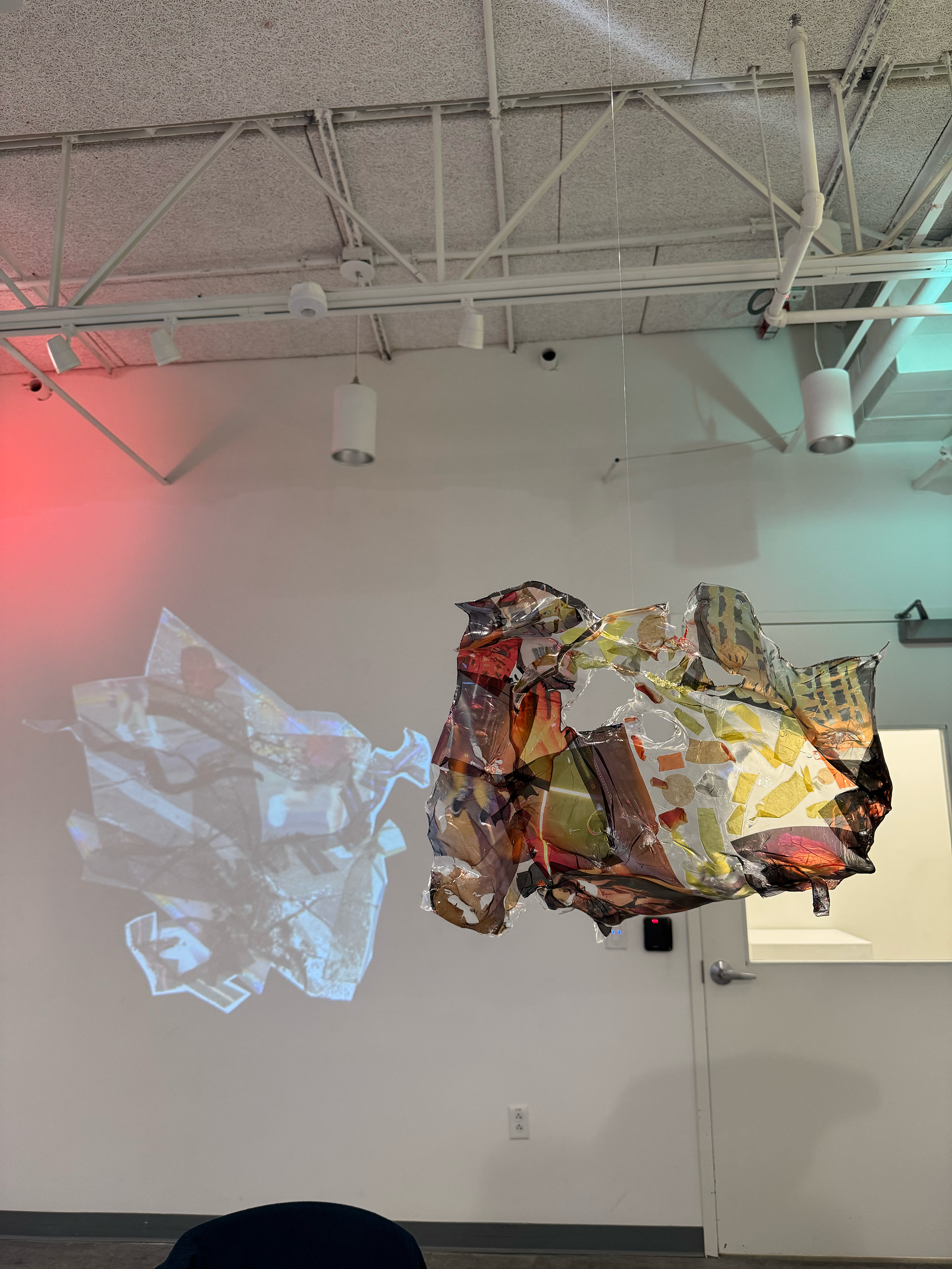 An abstract art sculpture made of crumpled and layered multicolored metallic and plastic materials hanging from the ceiling, with a light projection casting a shadow of the sculpture onto the plain white wall behind it.
