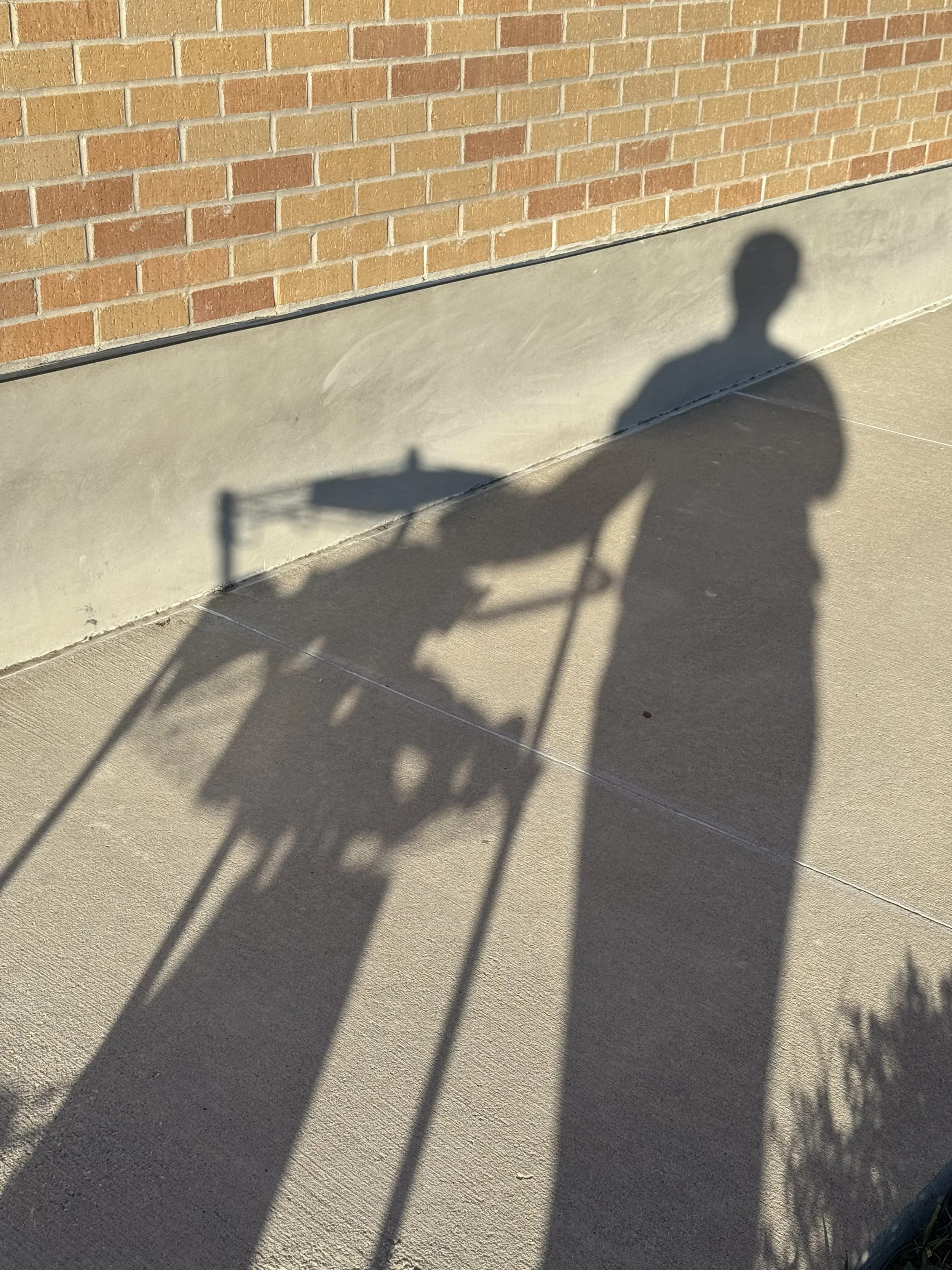 Shadow of a person pushing a stroller on a sidewalk with a brick wall in the background.