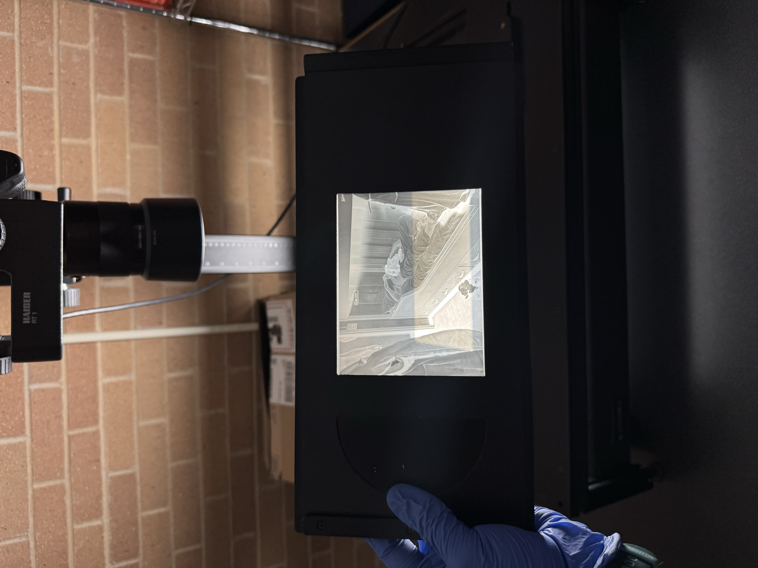 A person in blue gloves holds a glass slide over a black device with a mirror, in a lab setting against a brick wall.