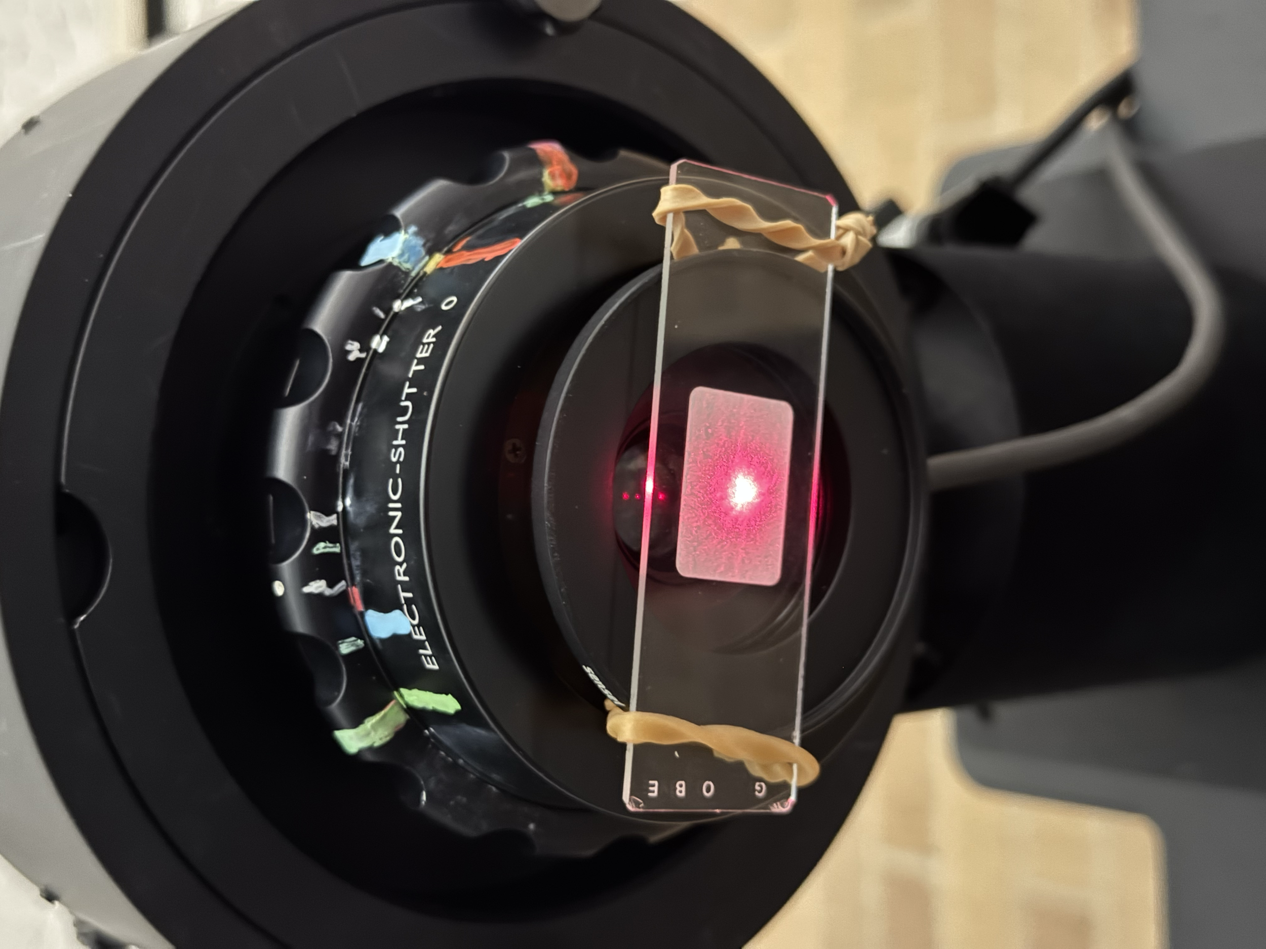 A laser device emitting a red beam through a glass and plastic setup, with various colored tape markings on the surrounding components.