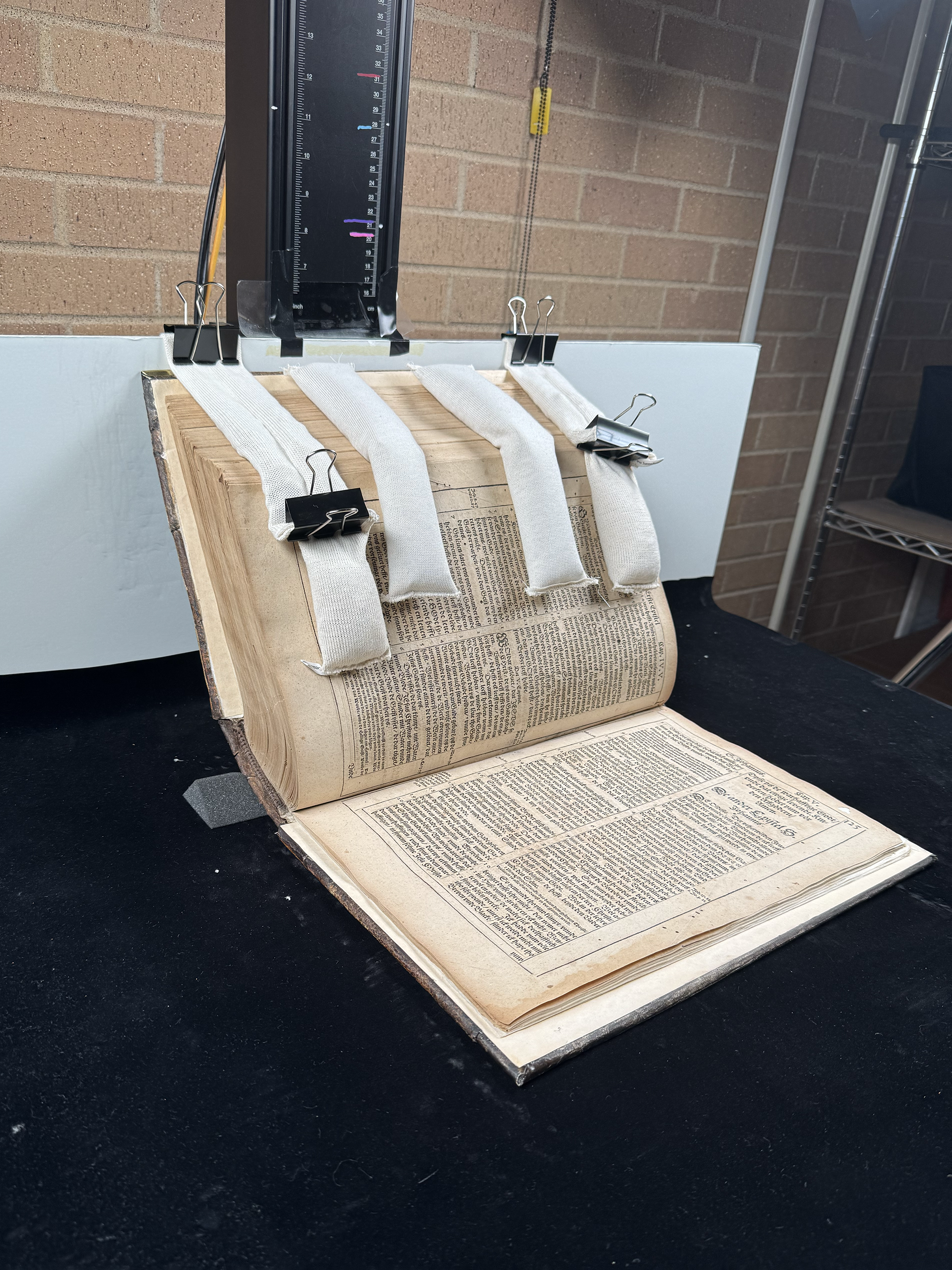 An old open book with fabric strips clipped to its pages, held in place by binder clips against a clipboard and a brick wall background.