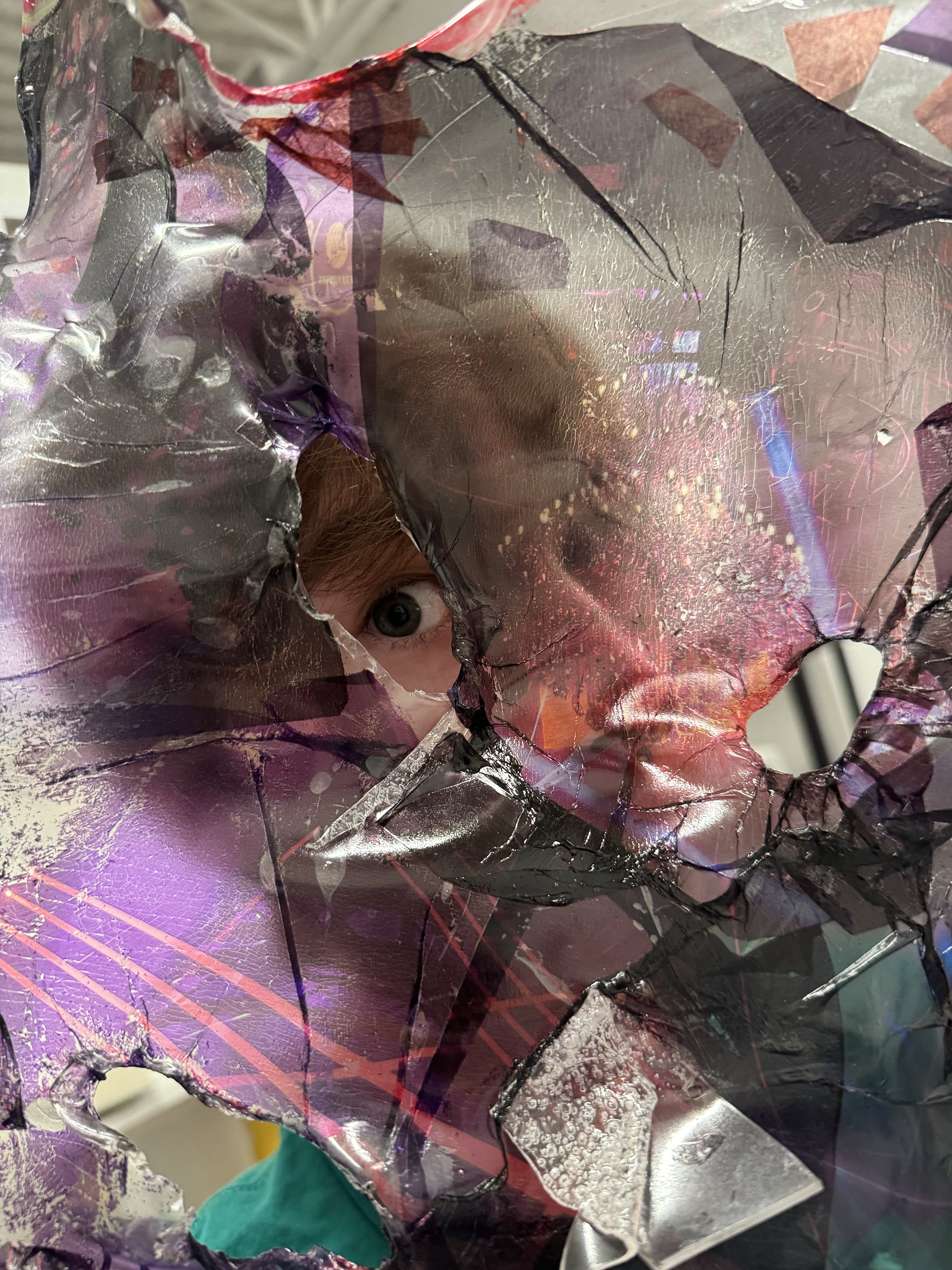 A person peeking through a large, irregular hole in a colorful, reflective, fractured plastic surface.