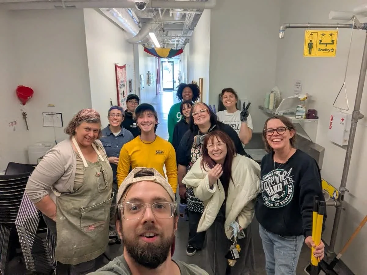 Group of people smiling and posing for a photo inside a building, with some making peace signs. The setting appears to be a kitchen or break room, with industrial pipes and medical equipment visible.