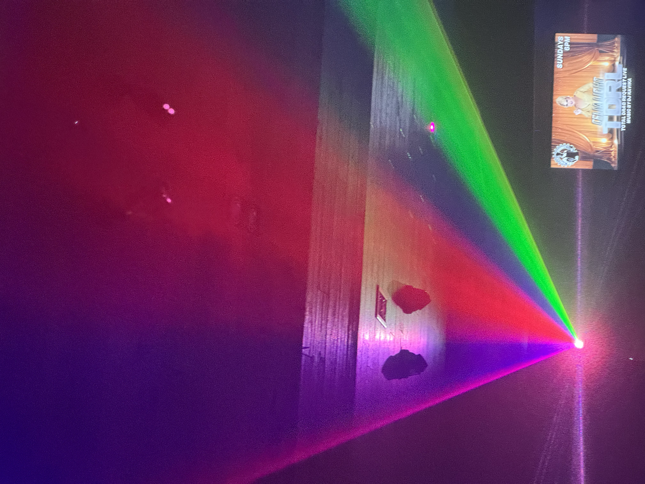 A dark room with multicolored laser lights projecting onto a wall and floor, and a promotional poster on the wall.
