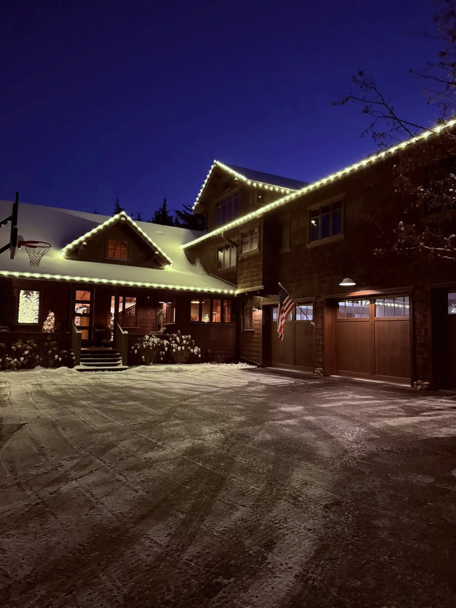 A house decorated with Christmas lights at night, with snow on the roof and driveway, American flag near the entrance.