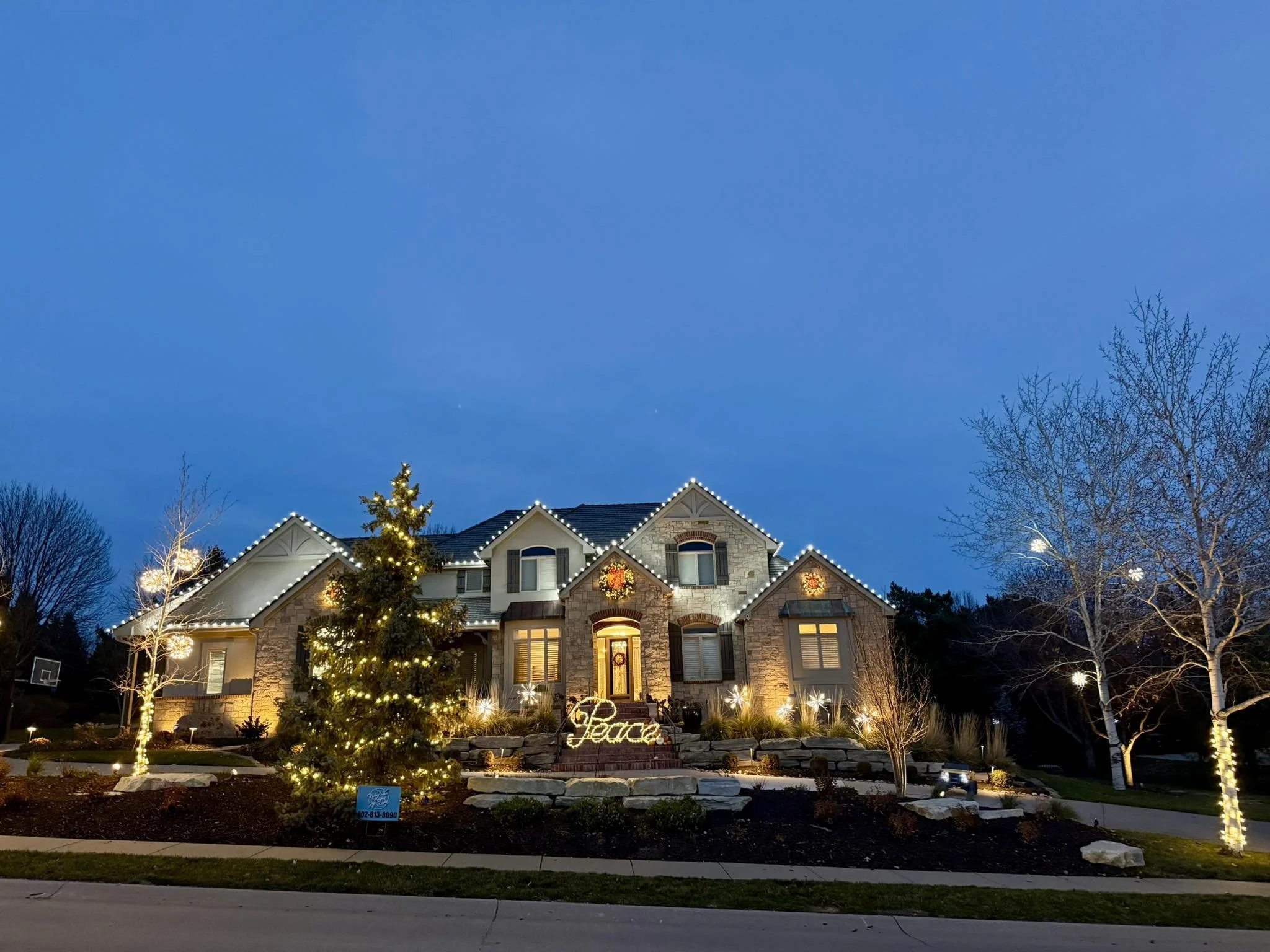 A large house decorated for Christmas with Christmas lights, wreaths, and a glowing sign that says 'Peace' in the front yard, surrounded by leafless trees with lights.